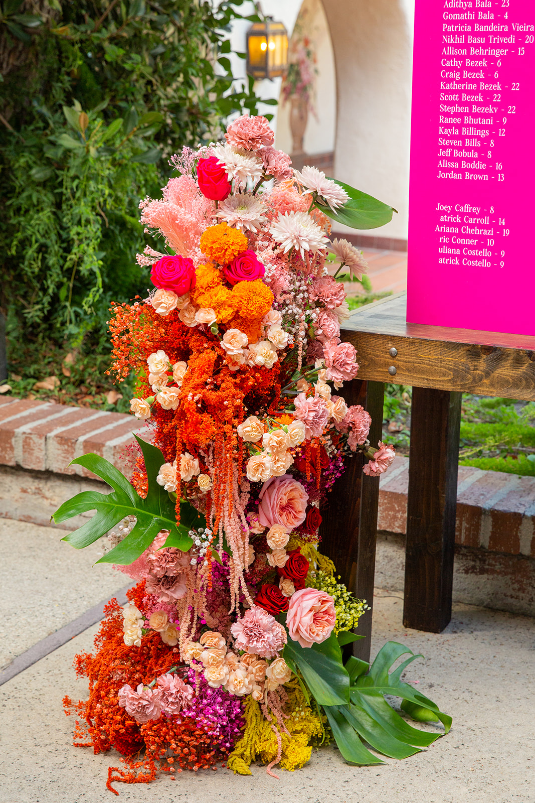 Still life shot of a large floral arrangement with a mix of roses, carnations, and various filler flowers in shades of pink, orange, and red, cascading down in an elaborate, almost waterfall-like manner. The flowers vary in size and type, creating a rich texture and a visually stunning impact. A bright pink seating chart sits on a table next to it, guiding people to their seats.