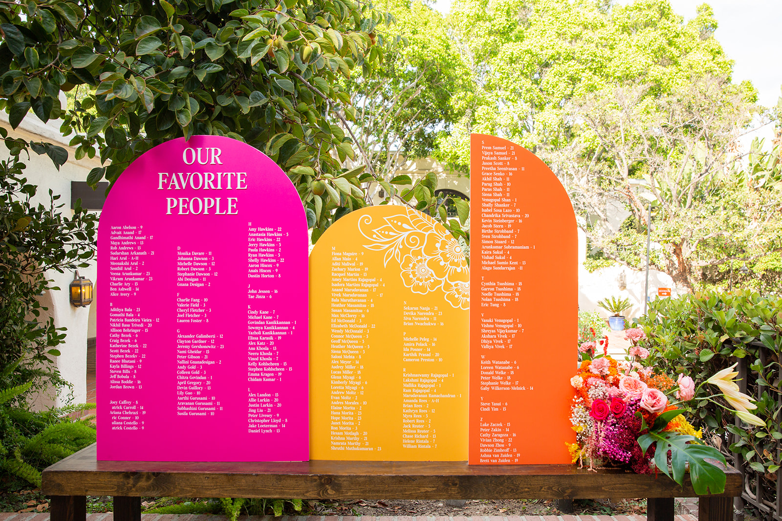 Wide shot of the seating chart. Three arched display boards set against a lush garden backdrop, titled "Our Favorite People." The boards are vividly colored in pink, yellow, and orange shades.