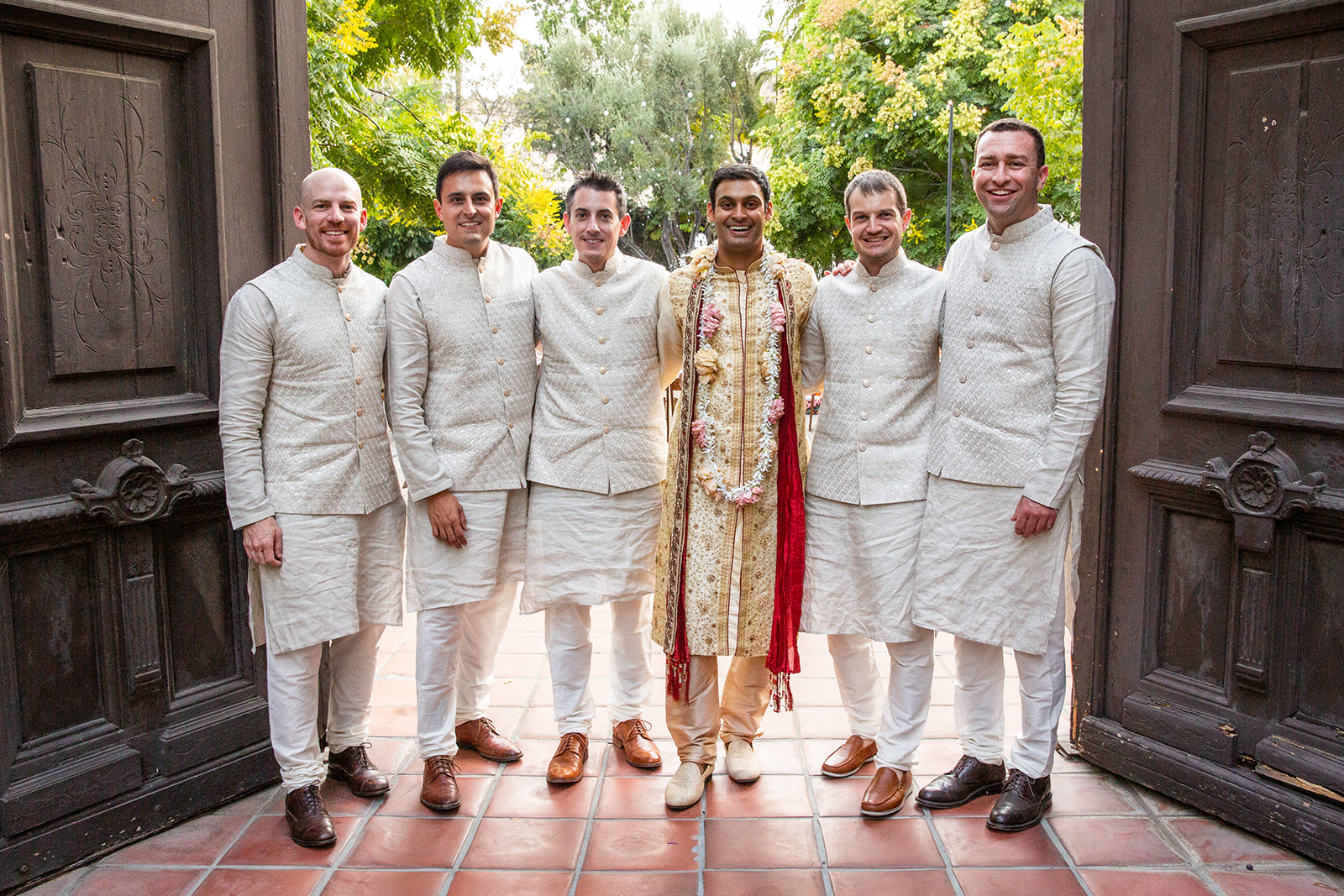 This wide shot features the groom and his groomsmen. The groom stands in the center, dressed in traditional Indian wedding attire, wearing a richly decorated cream and gold sherwani adorned with intricate embroidery. A red scarf draped over his shoulder adds a striking accent of color. Around his neck, he is wearing a garland of flowers. The groomsmen are all dressed in matching cream-colored outfits: knee-length kurtas with quilted, textured jackets. They stand under a large wooden archway or entrance, which frames the photo.
