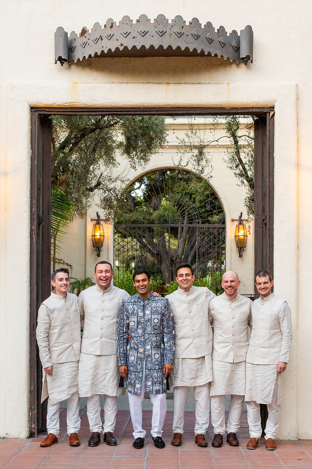 Wide shot of the groom, wearing a blue patterned outfit, standing in light cream-colored attire with his groomsmen. They’re all smiling and standing under a large archway with greenery in the background.