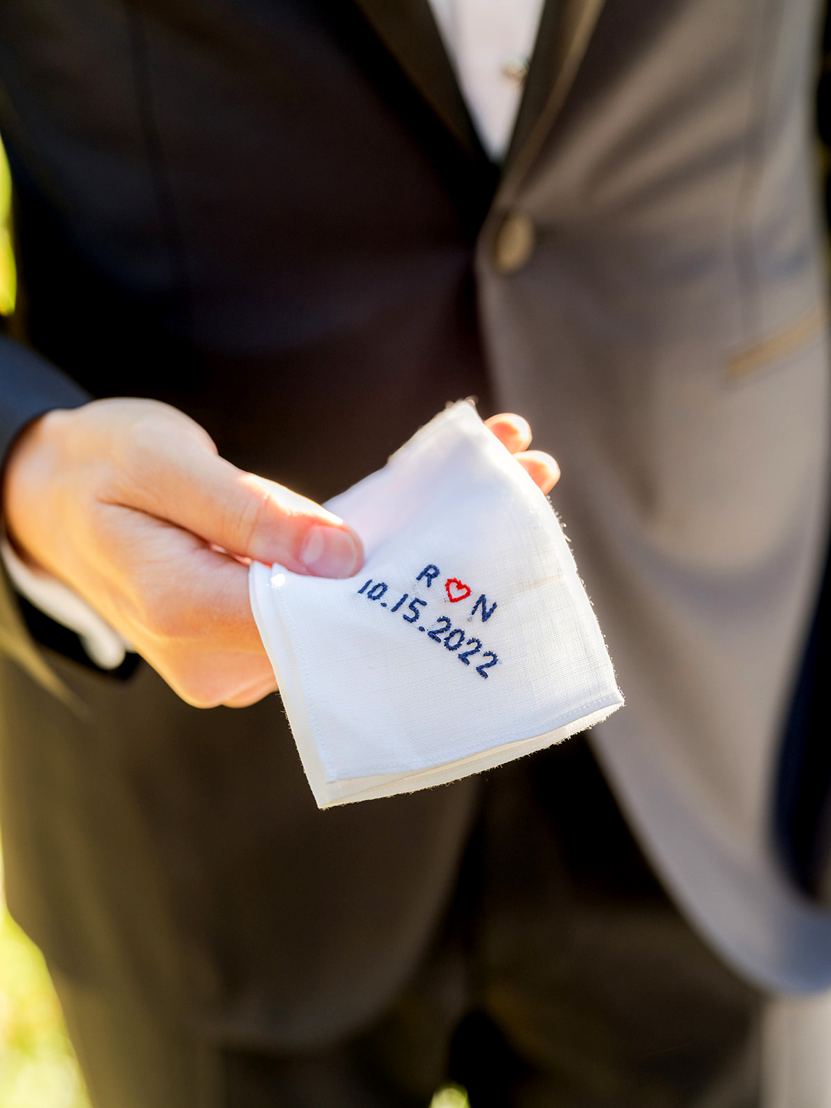 Close-up of the groom holding a small, white handkerchief personalized with embroidered text in blue that reads, "R ❤️ N" with the date "10.15.2022" beneath it. The "R" and "N" are separated by a small red heart, symbolizing the couple