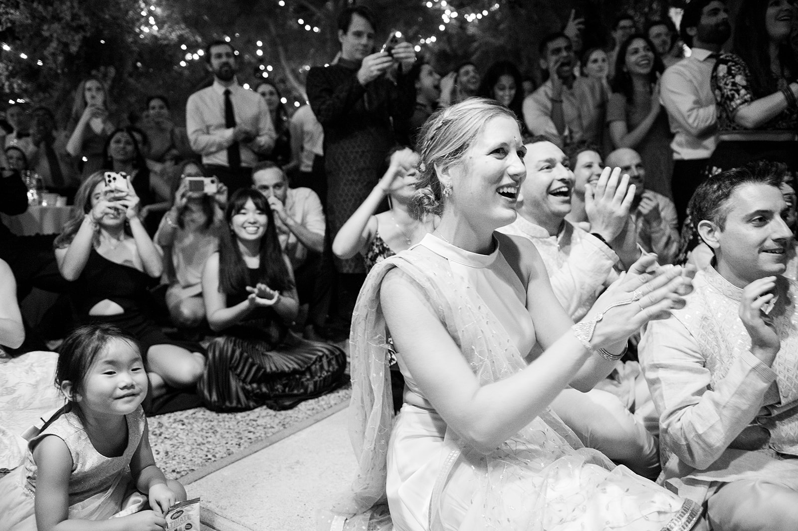 This black-and-white image captures a lively crowd. People are seated and standing, smiling, clapping, and taking phone photos. A woman in traditional attire, sitting in the front, looks particularly joyful, along with those around her. The background is adorned with string lights, and a young girl sits on the floor in front, smiling calmly and enjoying the event.