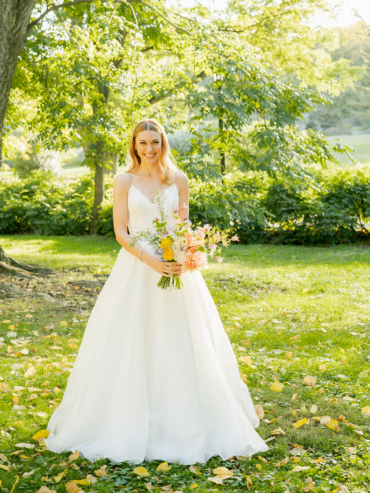 A wide shot of N standing outdoors in a lush, green park holding a bouquet includes various soft peach, yellow, and pink flowers. Yellow and green leaves are scattered on the grass beneath her. The background is filled with large trees and bushes, and sunlight filters through the leaves, casting a soft, natural glow over the scene.