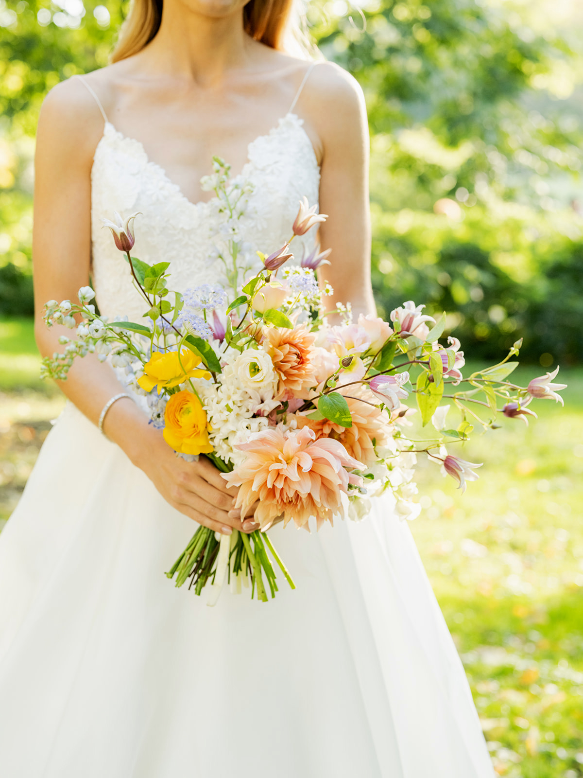 Close-up of our bride holding a colorful bouquet. The bouquet is a mix of soft peach, yellow, pink, and white flowers, including large dahlias and small wildflowers, with bright green leaves and stems adding a touch of freshness.