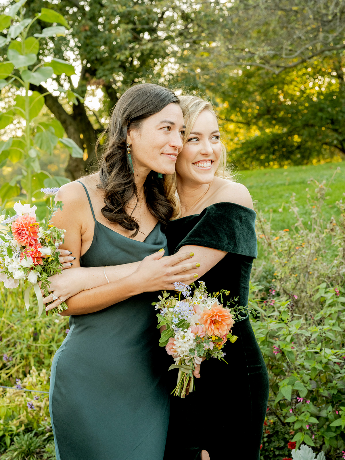 Medium shot of two bridesmaids embracing, smiling warmly in a garden setting. They are dressed in elegant, dark-colored dresses: one wears a deep green satin dress, while the other wears a dark velvet off-the-shoulder gown. Both hold small bouquets of colorful flowers with bright orange dahlias, white blooms, and green foliage.
