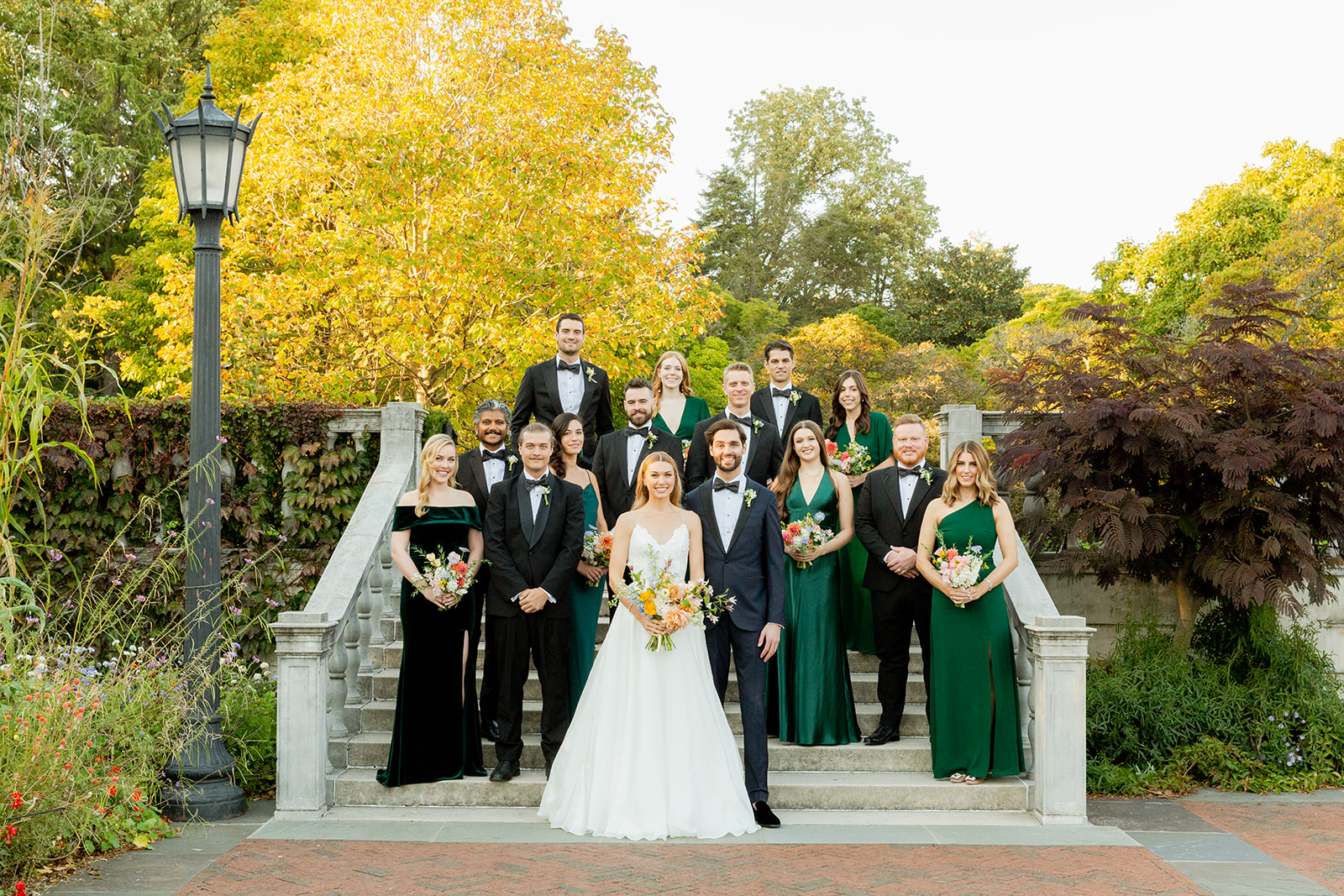 Wide shot of a formal wedding party portrait. The bride and groom are at the center, standing on a set of outdoor stone steps, surrounded by their bridesmaids and groomsmen. The bridesmaids wear elegant green dresses, and the groomsmen wear black tuxedos. Each bridesmaid holds a bouquet with bright orange, pink, and white flowers, adding vibrant pops of color against the green and black outfits. The group is arranged symmetrically, with the bride and groom at the front and the rest standing on the steps behind them.