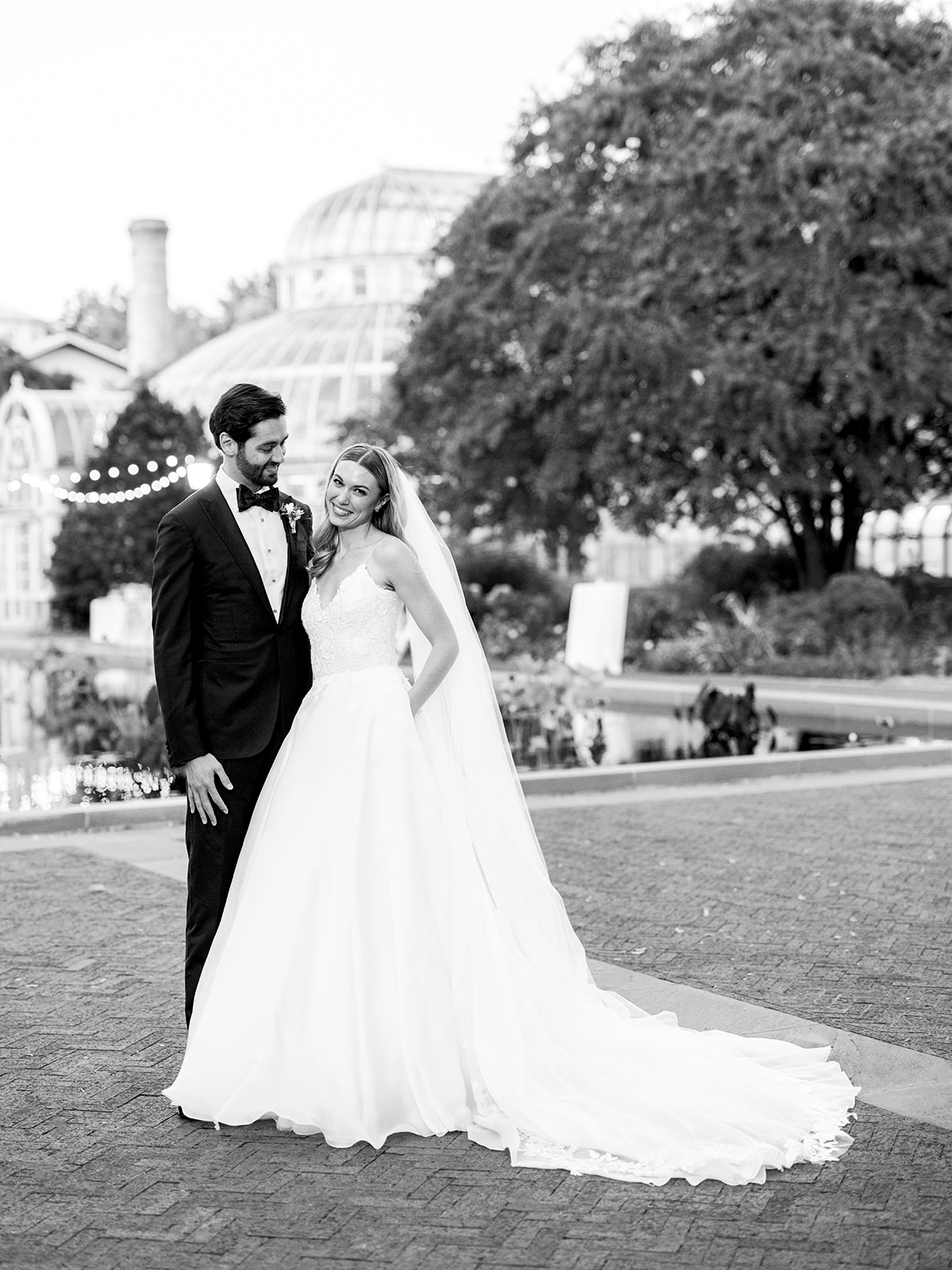Similar shot of the bride and groom in black and white in the early evening standing together outside of the wedding reception. The bride smiles at the camera as the groom gazes at his bride.
