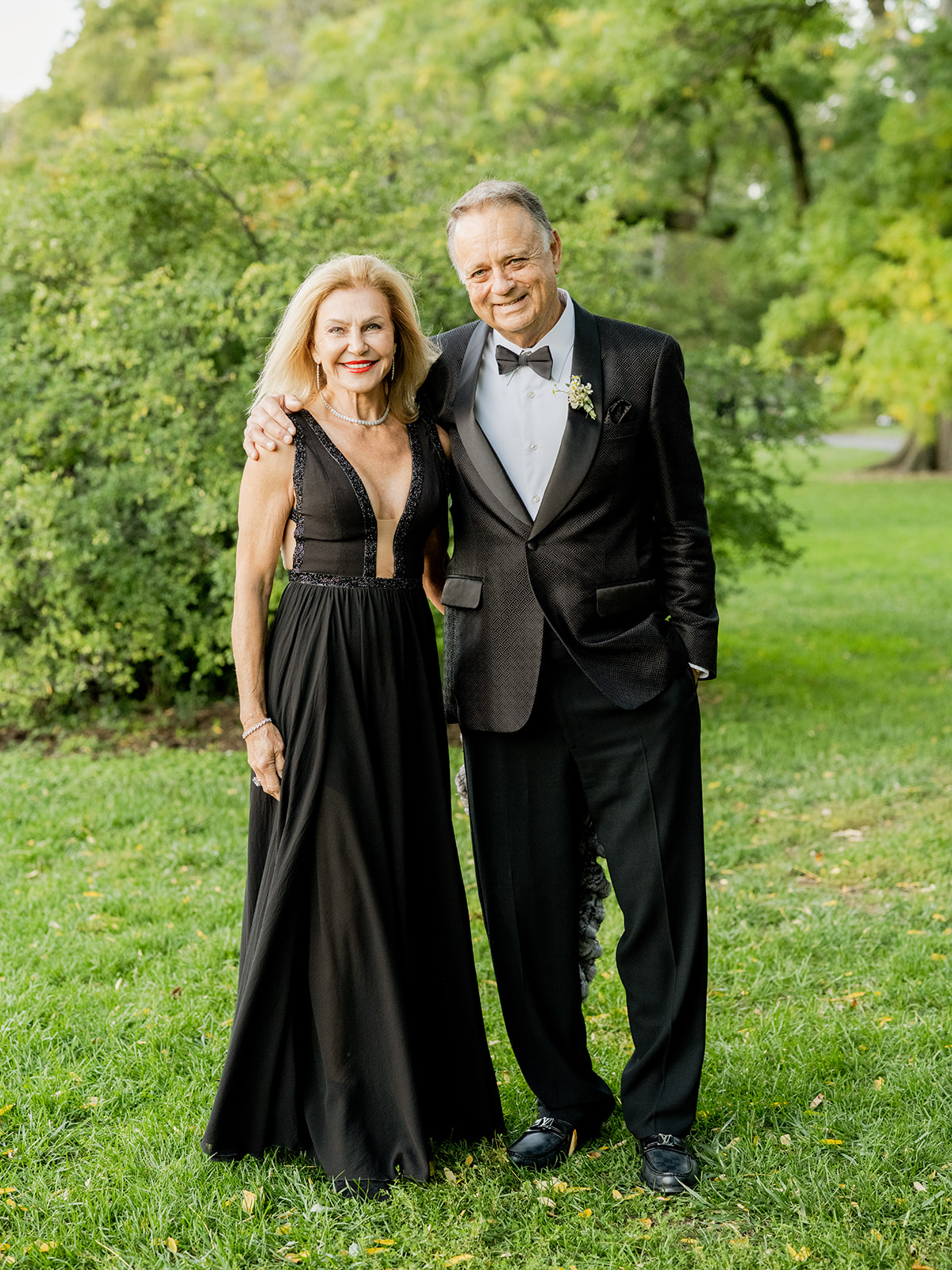 Wide shot of a couple standing outdoors in the lush green park. He wears a formal black tuxedo with a white shirt and black bow tie, complemented by a small boutonniere on his lapel. He has his arm around the woman, smiling warmly at the camera. The woman is wearing a sleeveless, long black gown with intricate details, including a deep V-neckline and lace accents. She smiles confidently, with bright red lipstick adding a pop of color.