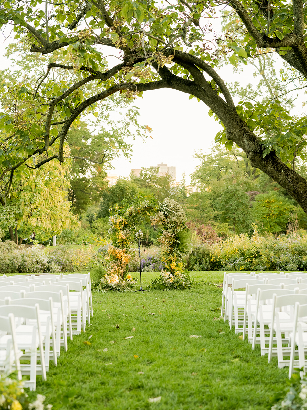 Wide shot of a beautifully decorated floral arch made of greenery and soft yellow and white flowers, positioned at the end of a grassy aisle. On either side of the aisle, rows of white folding chairs are neatly arranged and ready for guests. A large, overarching tree frames the scene, with its branches spreading overhead, adding a natural canopy to the ceremony space.
