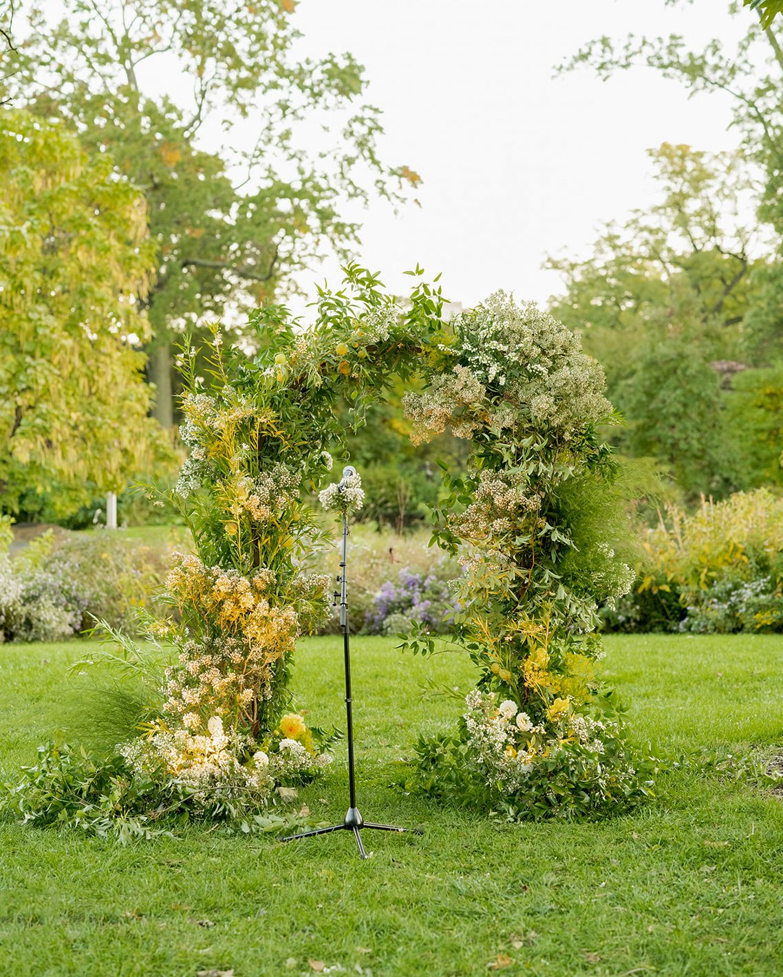 Close-up shot of the floral arch made of greenery and soft yellow and white flowers. In the background, more greenery and trees fill the space.