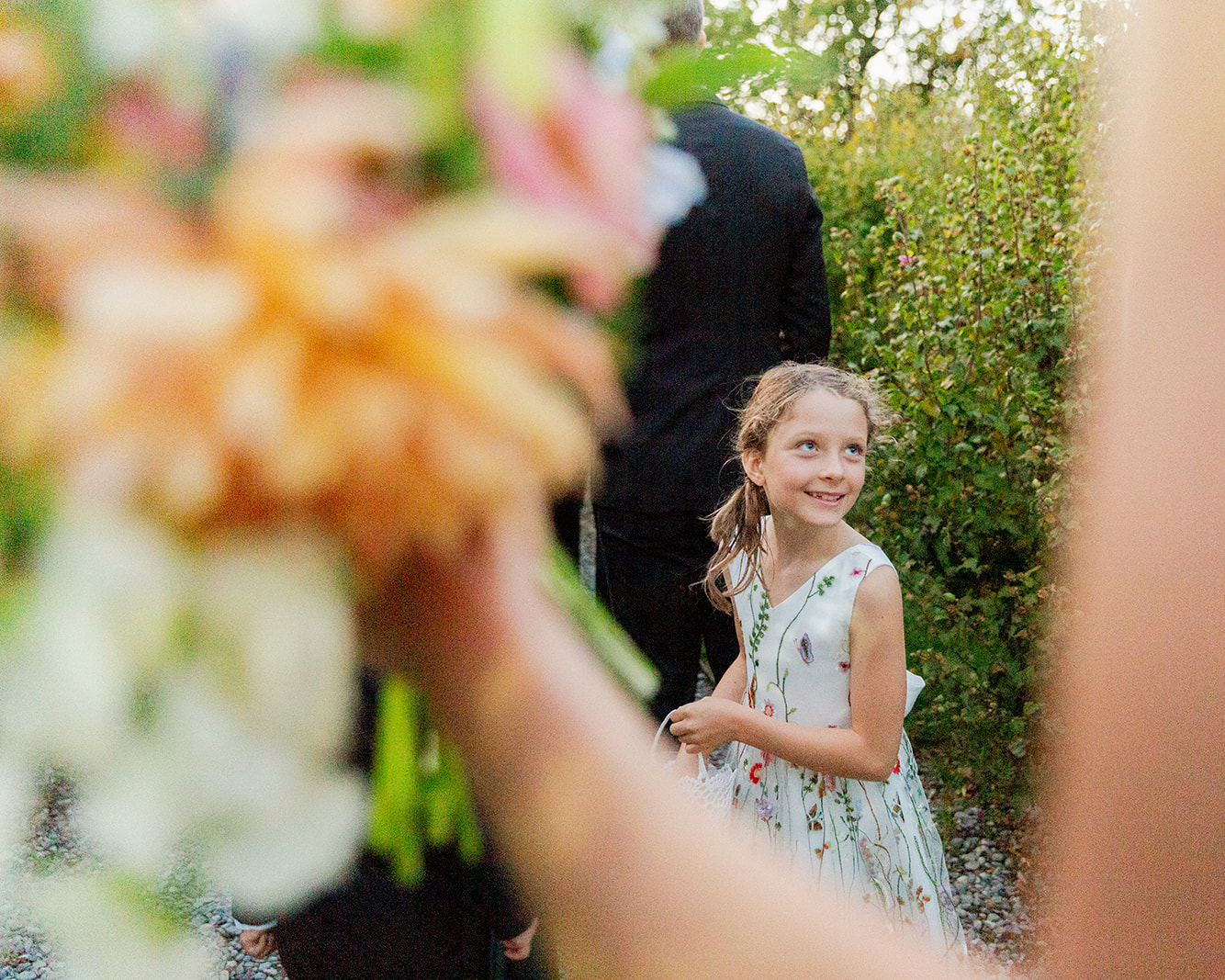 Candid moment of the flower girl smiling warmly, looking off to the side with a joyful expression. She is wearing a light-colored, floral-patterned dress. Her hair is pulled back into a ponytail. In the foreground is a large, out-of-focus bouquet, mostly orange and white, which slightly obscures part of the image.