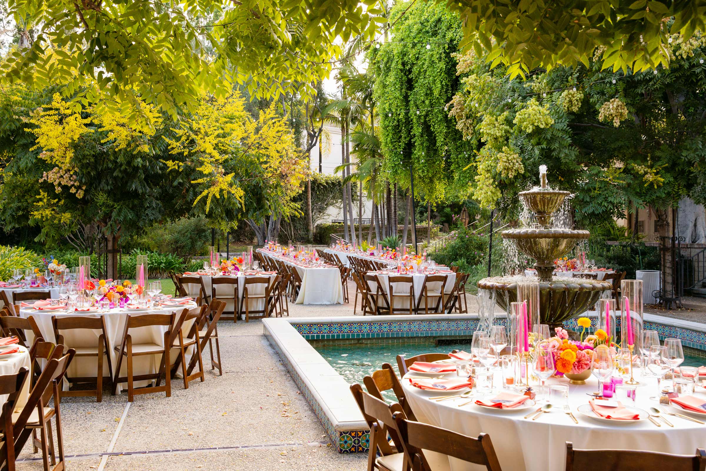 Wide shot of circular tables dressed in crisp white tablecloths neatly arranged around a central decorative fountain. Each table is festively adorned with vibrant pink and orange floral centerpieces, complemented by matching place settings and tall glass candle holders. The scene is surrounded by mature trees and dense foliage, with dappled sunlight filtering through the leaves.