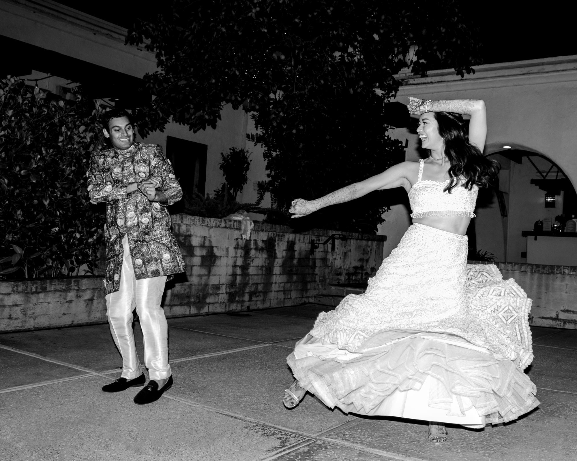 This black-and-white image captures a joyful moment between the bride and groom dancing. The groom, dressed in a detailed, patterned sherwani, looks happy and playful as he moves. The bride, wearing a flowing lehenga, is twirling, with her skirt flaring out as she spins. Her arms are raised in an expressive, joyful gesture, and her face shows pure happiness.