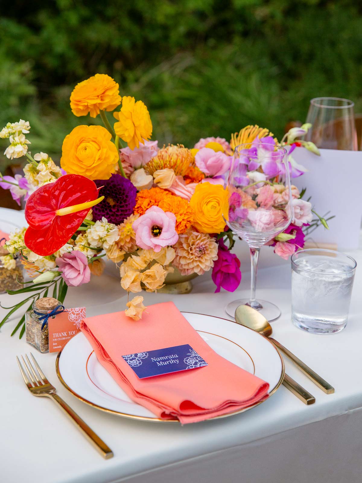 The image shows a close-up of a beautifully arranged table setting with bright and vibrant colors. A plate with a coral-colored napkin is at the center of the image. On top of the napkin, there