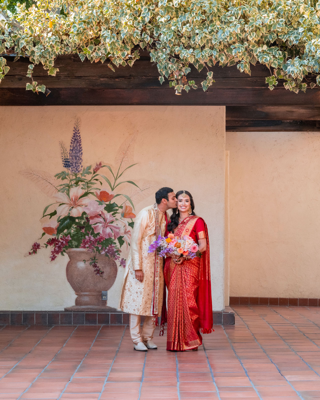 Full body shot of A and H as they stand together under a rustic archway, enveloped by a canopy of green vines. A wears a traditional red and gold saree and holds a bouquet of vibrant flowers. At the same time, H complements her perfectly in a cream and gold traditional outfit that echoes the warm, sunlit environment. The backdrop features a large ceramic vase with an arrangement of tall flowers.