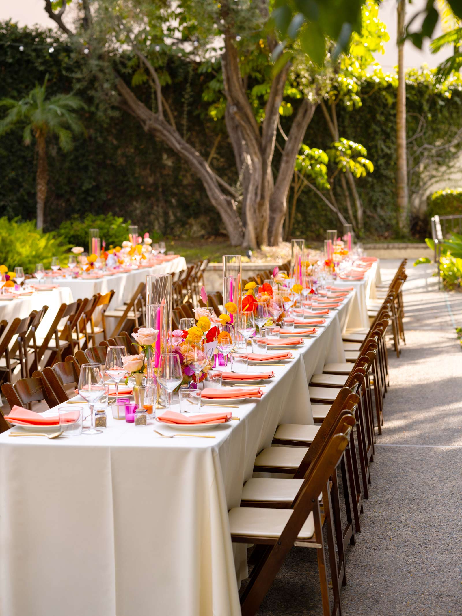 Wide shot of long tables adorned with ivory tablecloths and meticulously set with vibrant orange and pink accents, from the napkins to the glassware. The centerpieces are a colorful mix of various flowers, adding a rich texture and festive atmosphere to the setting.