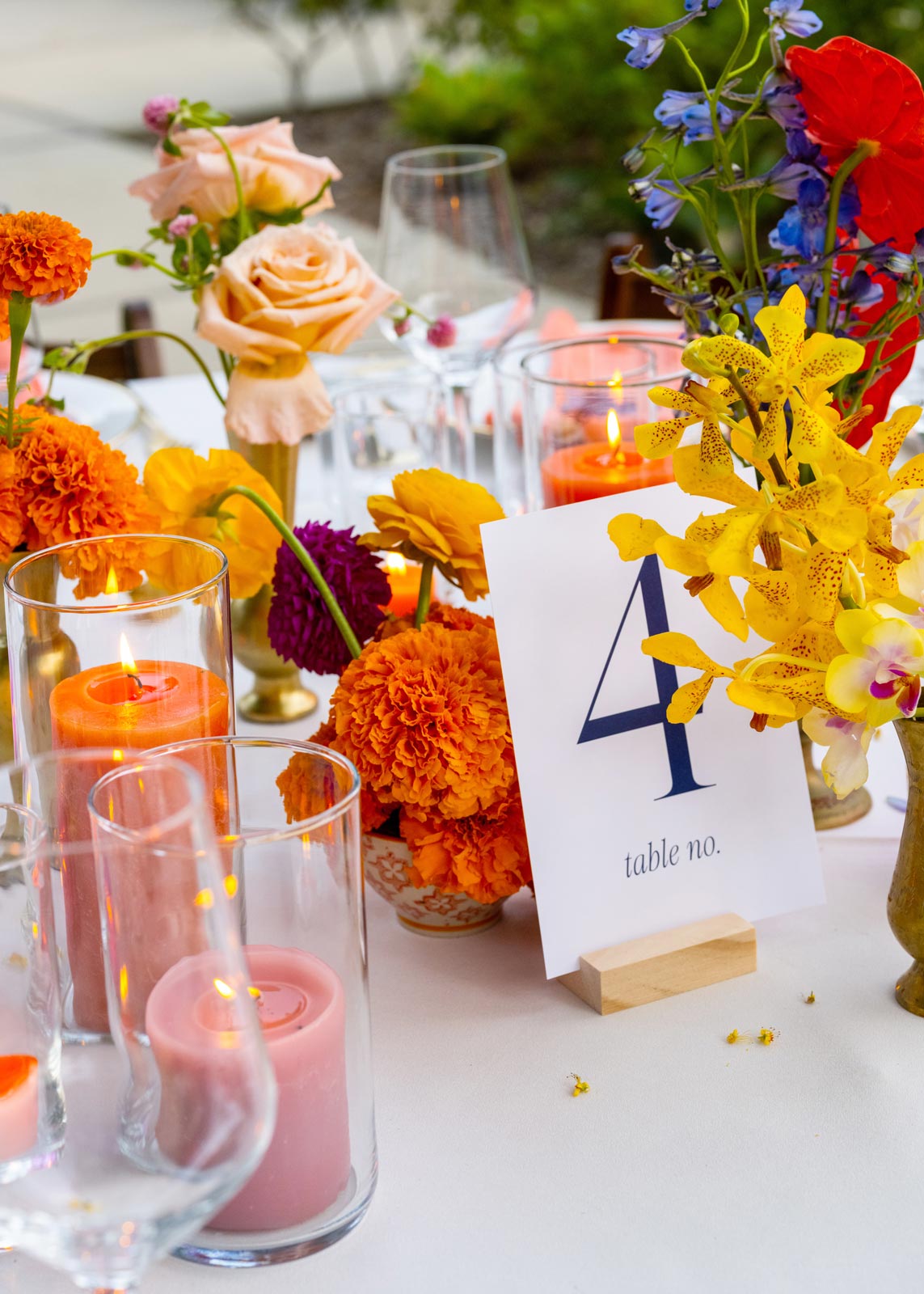 Another colorful close-up shot of the table centerpieces. This table is labeled "Table No. 4," with the table number displayed on a simple white card held in a wooden base. The typography on the card is clean and modern, with the number "4" in large, bold blue text. The centerpiece is composed of vibrant and colorful flowers in orange, yellow, and pink shades. Prominent flowers include bright orange marigolds, soft peach roses, and yellow orchids with delicate spotted petals. A few taller flowers, like deep blue delphiniums and a bold red anthurium, add height and contrast to the arrangement. The floral display is housed in a mix of gold vases, adding a touch of vintage elegance to the table decor. Surrounding the flowers are several glass votives holding candles in shades of coral and orange, creating a warm, intimate glow. The candles are lit, giving the scene a soft, inviting ambiance that complements the cheerful and vibrant colors of the flowers. Wine glasses are arranged around the table, catching the candlelight and adding a refined sparkle to the setting.