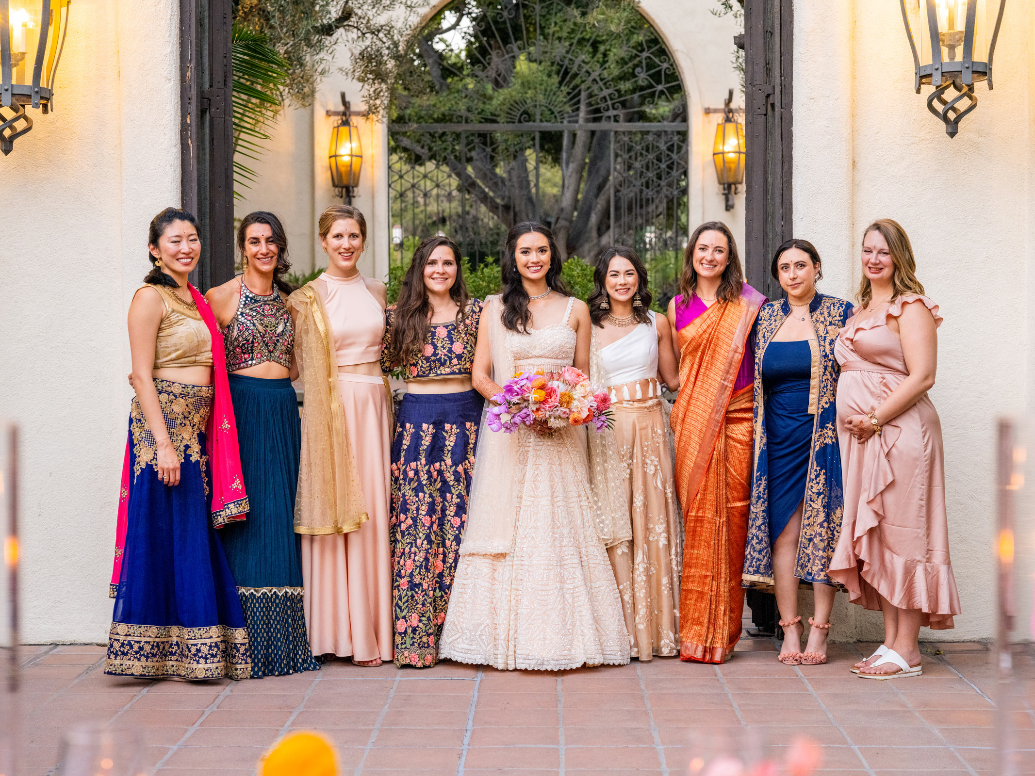 Wide shot of the bride standing with her bridesmaids, all smiling. The group is dressed in colorful, elegant outfits, including traditional Indian attire and modern dresses. They are standing outdoors under a large archway.