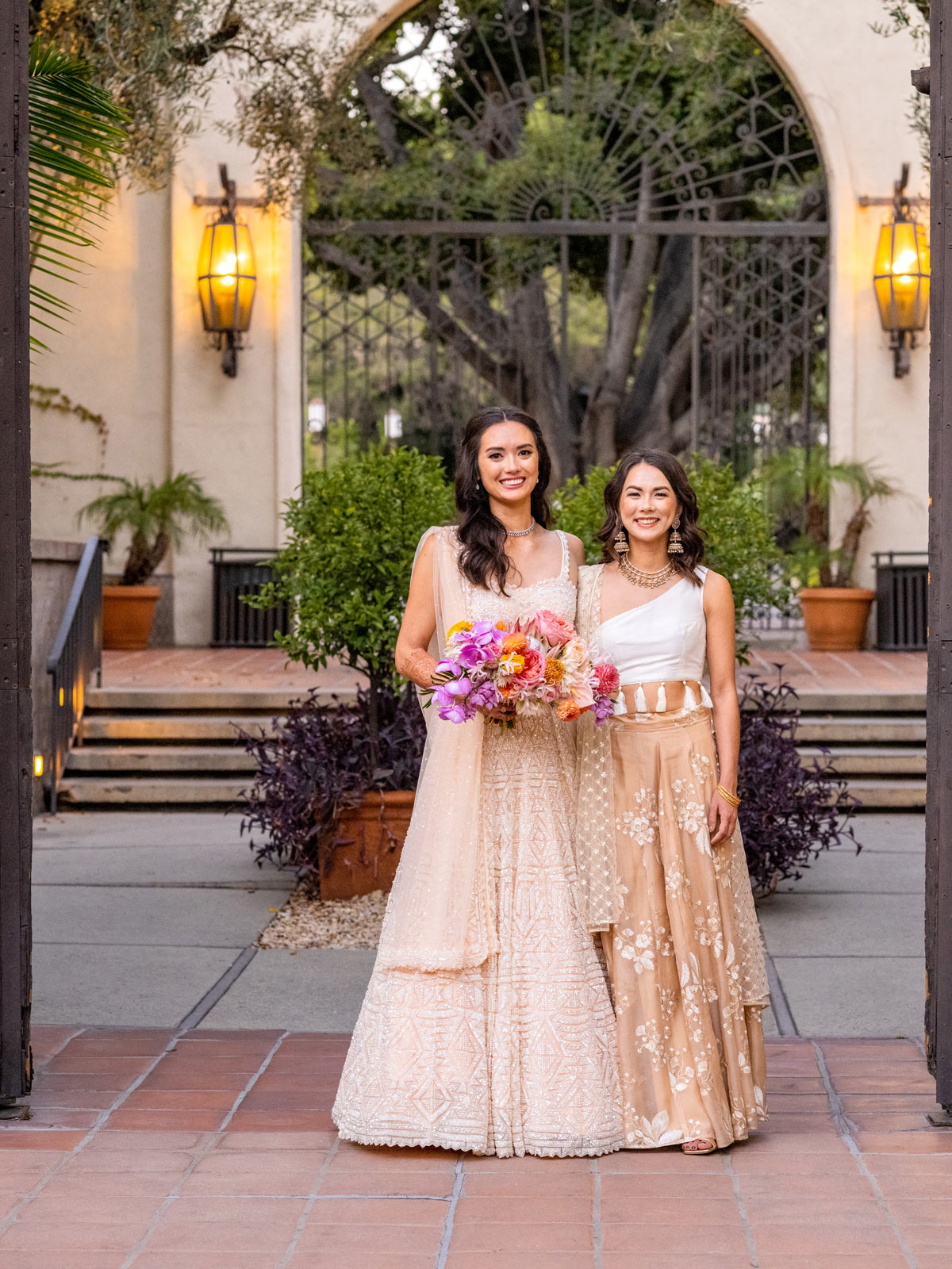 Wide shot of our bride, dressed in all pink with beautiful embroidery, standing next to her sister. The bride holds a colorful bouquet, and both women are smiling warmly. They’re outside in front of an arched doorway, with plants and soft lighting around them.