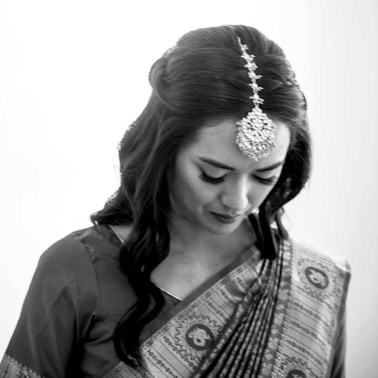 Medium black and white shot of A adorning a piece of jewelry on her head, a traditional piece that catches the light, highlighting intricate patterns and sparkling embellishments. The moment is reflective and quiet.