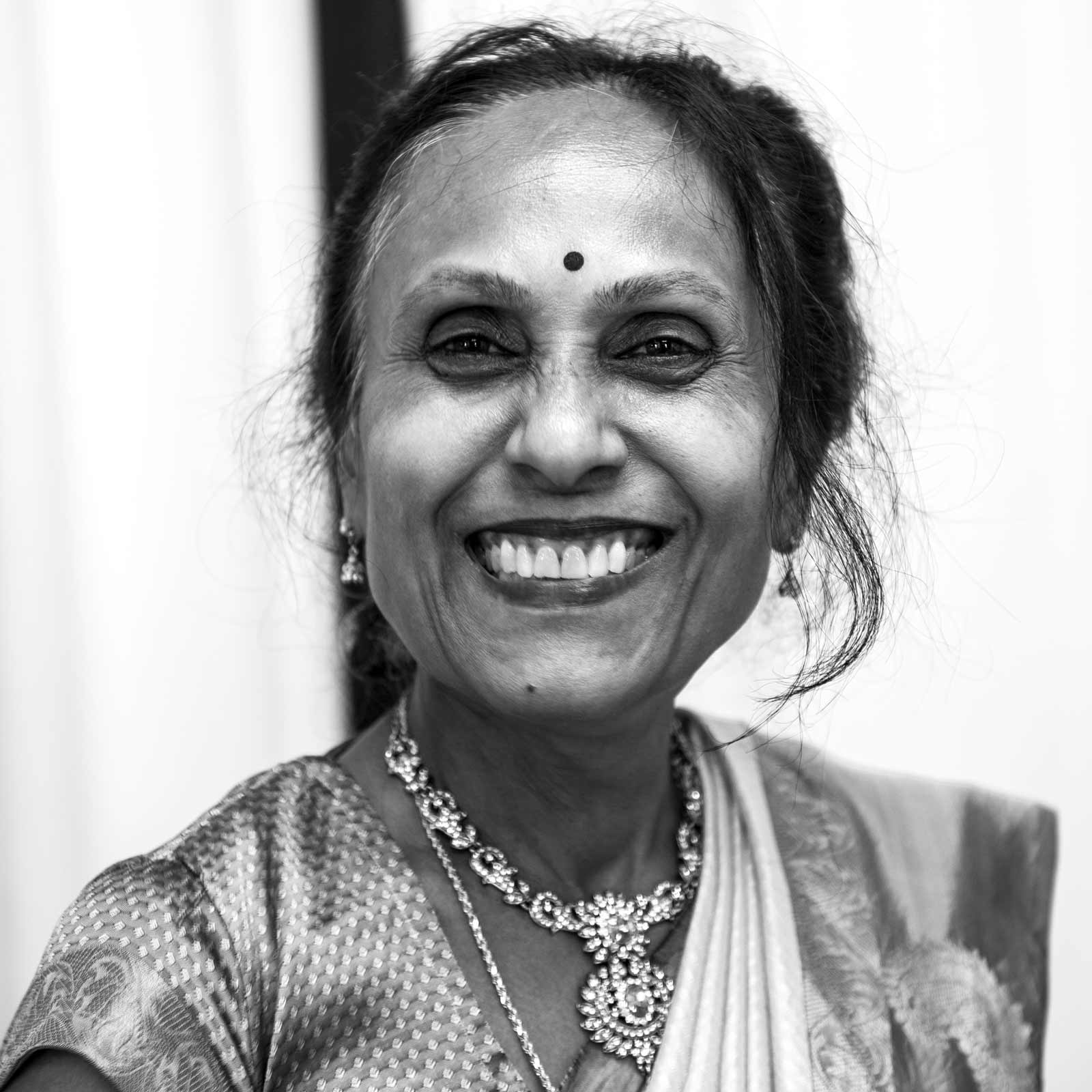 This black-and-white close-up portrait of H’s mom is expressive, radiating happiness, emphasized by crinkles around her eyes and a slight dimple on her cheek. She wears traditional attire, including a finely textured saree draped elegantly over her shoulder. A prominent, detailed necklace adorns her neck. Her hair is pulled back, and she sports a small, simple bindi on her forehead, a traditional mark worn often for cultural and spiritual reasons.