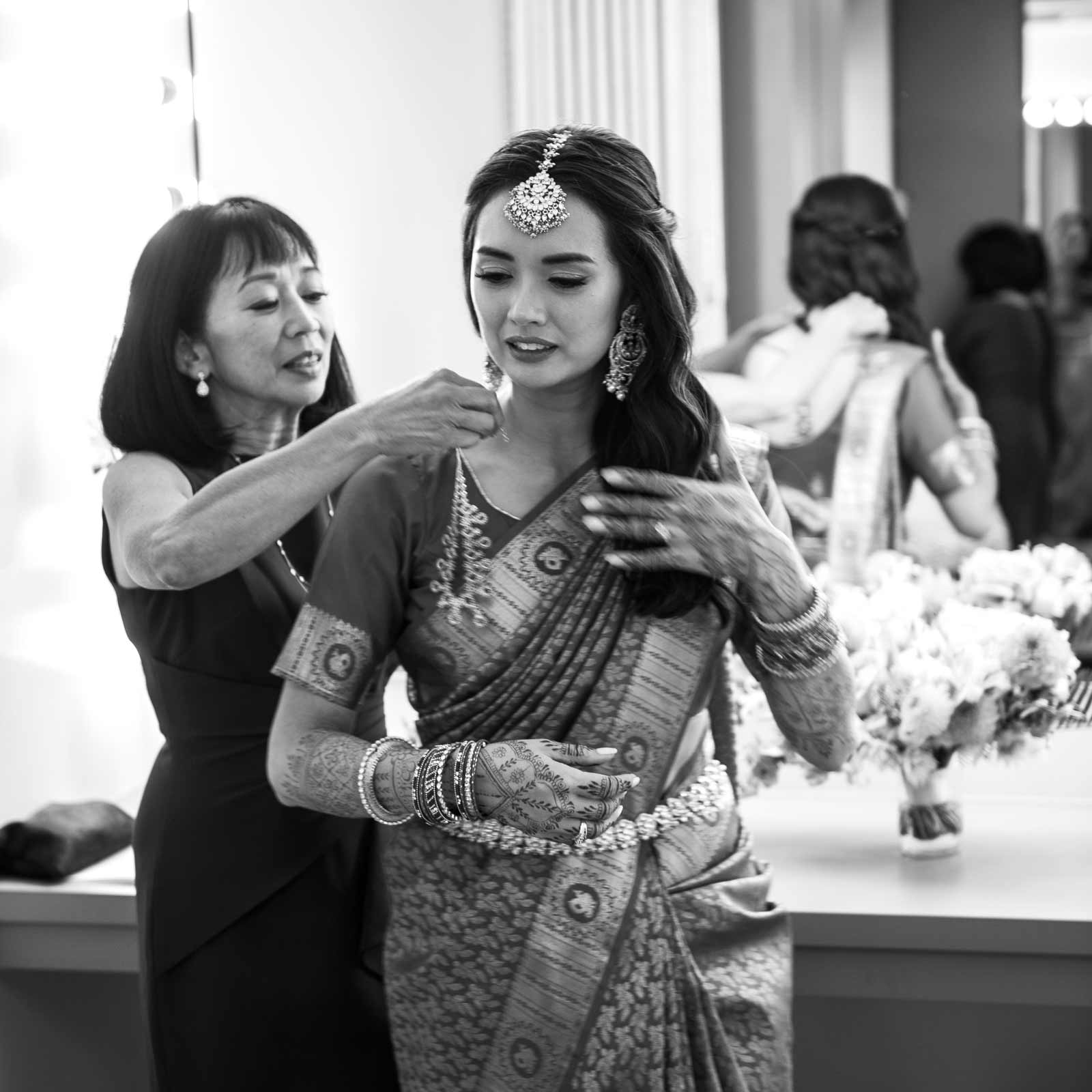This black-and-white medium shot of A with her mother captures a tender moment of preparation for the wedding. A is dressed in traditional attire, her saree intricately patterned, paired with a fine set of jewelry that includes large earrings and a headpiece. Her hands are decorated with detailed henna designs, and she wears numerous bangles. They are in a brightly lit room with large mirrors lined with bulbs, reflecting multiple images of the activity and enhancing the intimate, busy atmosphere.
