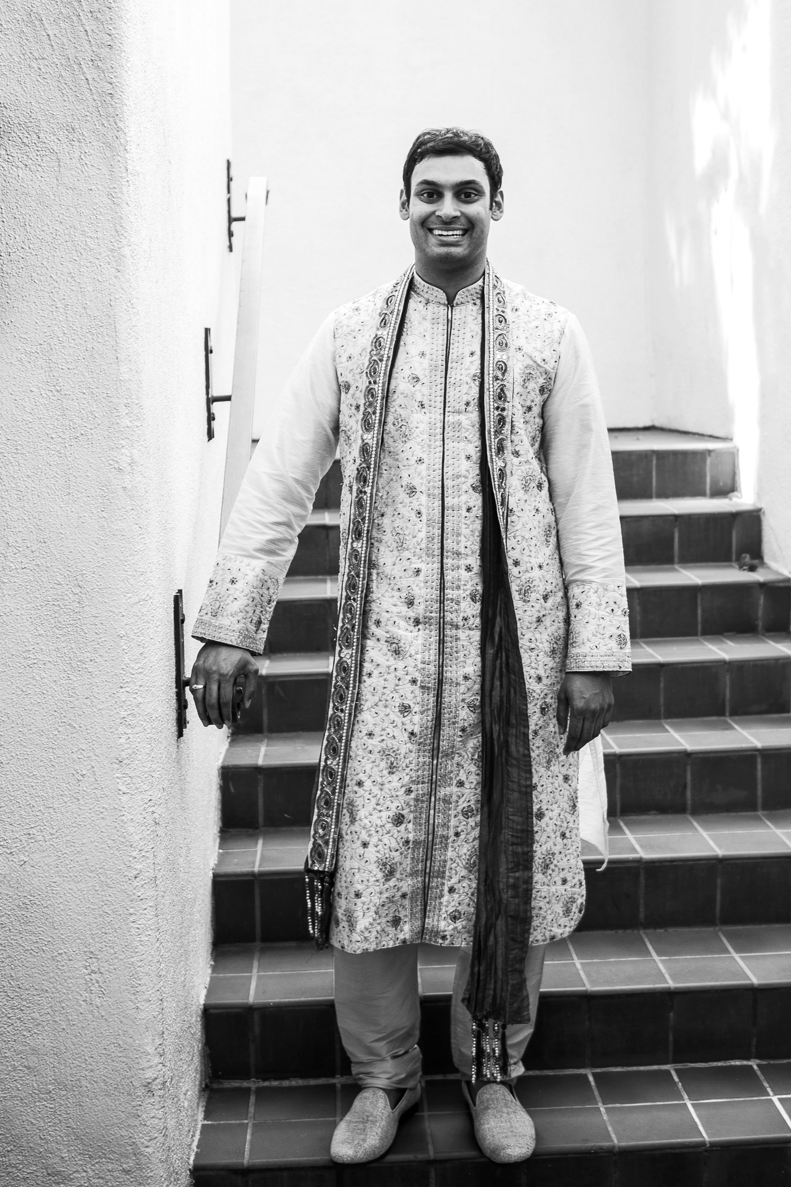 Wide black-and-white shot of our groom H as he stands on a staircase waiting to see his bride A for the first time of the day. He is dressed in traditional Indian attire, which includes a long, ornate kurta with intricate designs and a matching scarf draped over one shoulder.