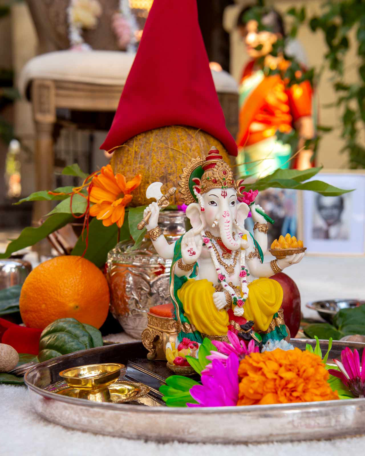 Close-up shot of a statue of Lord Ganesha, an important deity in Hinduism known as the remover of obstacles and the god of beginnings. The statue is intricately painted and adorned with jewelry, positioned in the center of a ceremonial setup that includes various ritual items. Ganesha is seated, holding a small axe in one hand and a bowl of sweets in another, symbolizing rewards for wise and virtuous living. Surrounding the statue are vibrant offerings and elements used in Hindu worship: fresh flowers in bright pinks and oranges, fruits like oranges, and coconut, a traditional offering symbolizing purity and the ego that must be broken to reveal the sweetness within.