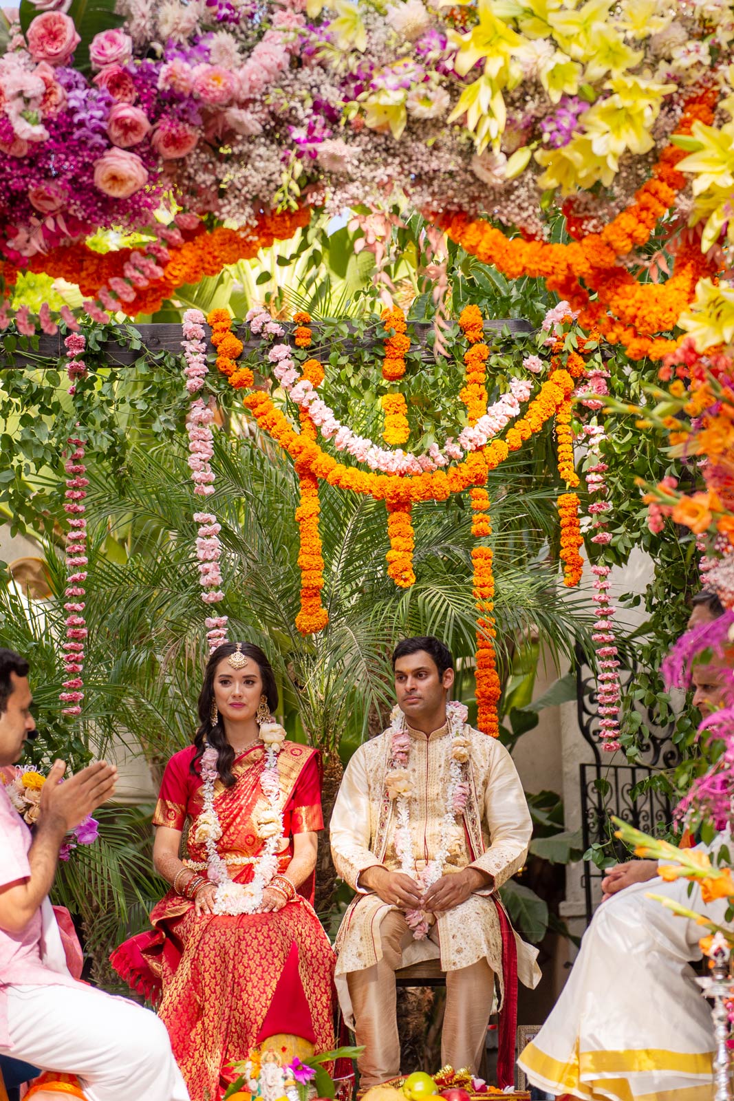 A wide shot of the bride and groom seated under the floral mandap. The couple is surrounded by a breathtaking array of flowers in vibrant colors, creating a stunning visual feast. Above them, the canopy is densely packed with blooms in shades of orange, pink, purple, and yellow, interspersed with lush green foliage. In front of them, a priest in a pink shirt conducts the ceremony, speaking and guiding the rituals, which include various offerings visible on the altar before the couple. These offerings typically consist of fruits, flowers, and other items significant to Hindu rituals, symbolizing prosperity, fertility, and purity.