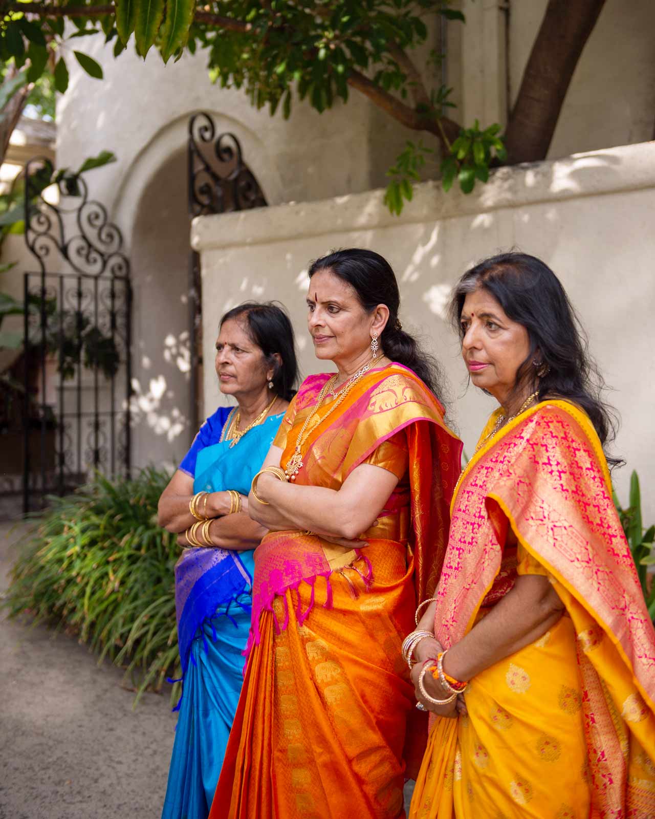 Medium shot of three women dressed in traditional Indian sarees, each vibrant in color and rich in detail. From left to right, the sarees are in bold blue, deep orange, and bright yellow, embellished with intricate patterns that glimmer subtly in the natural light.
