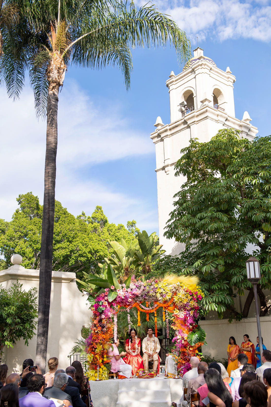 An even wider shot of the bride and groom seated under the floral mandap with the bell tower of the gardens in the background.