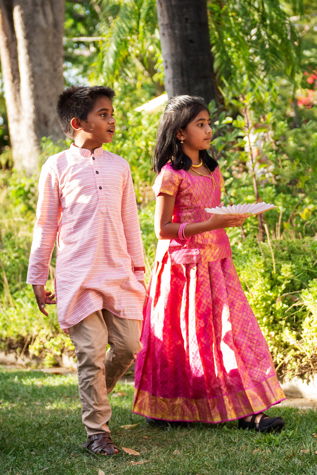 Wide shot of two young children walking through a lush garden of the ceremony. The kids dressed in traditional Indian attire. The boy is in a pale pink kurta with a striped pattern, and the girl is in a striking pink and gold lehenga, which is richly patterned and festive.
