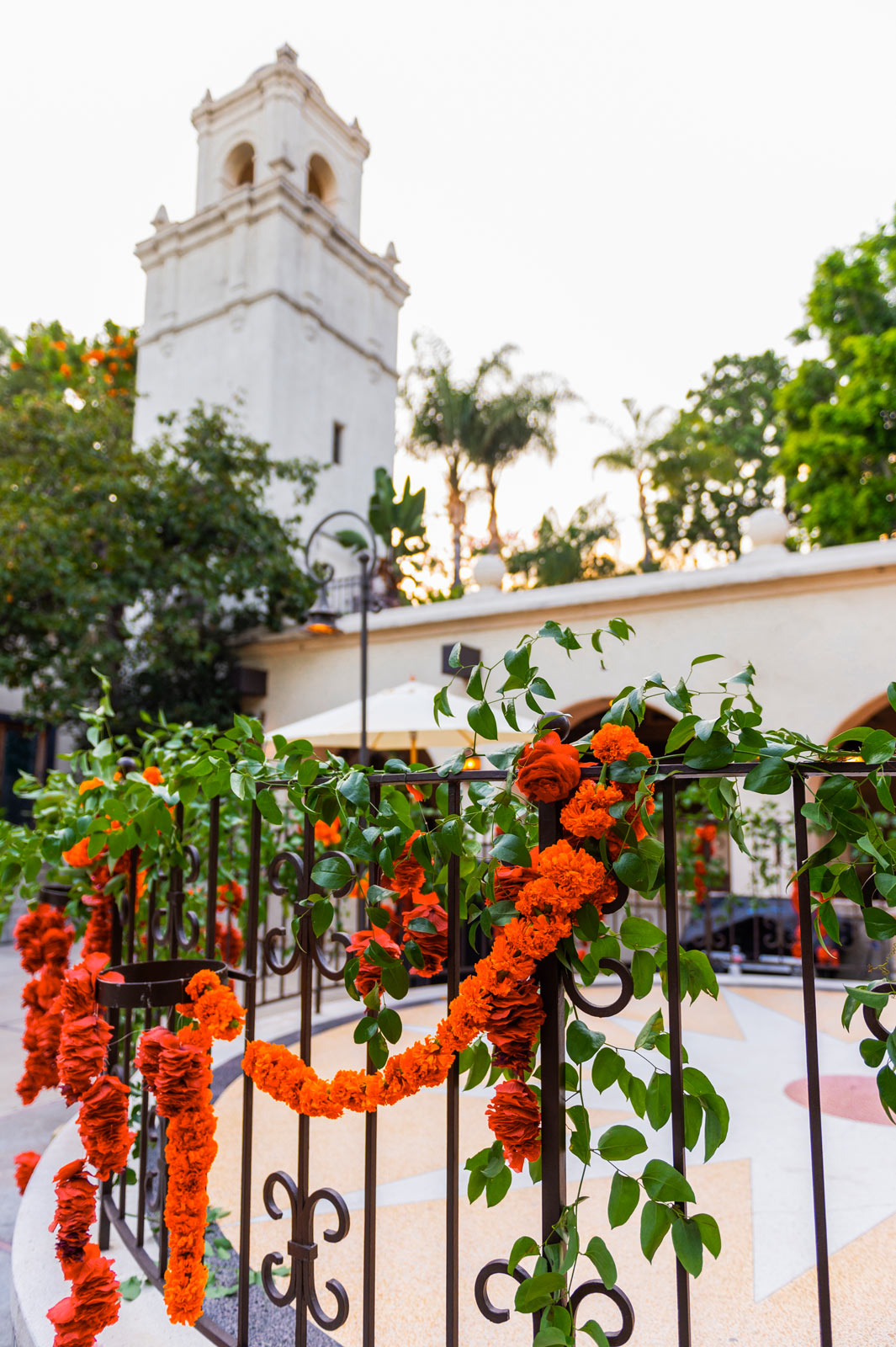 This wide shot image shows a tall, white bell tower in the background, with a decorative wrought iron fence in the foreground. The fence is adorned with garlands of red and orange flowers intertwined with green leaves.