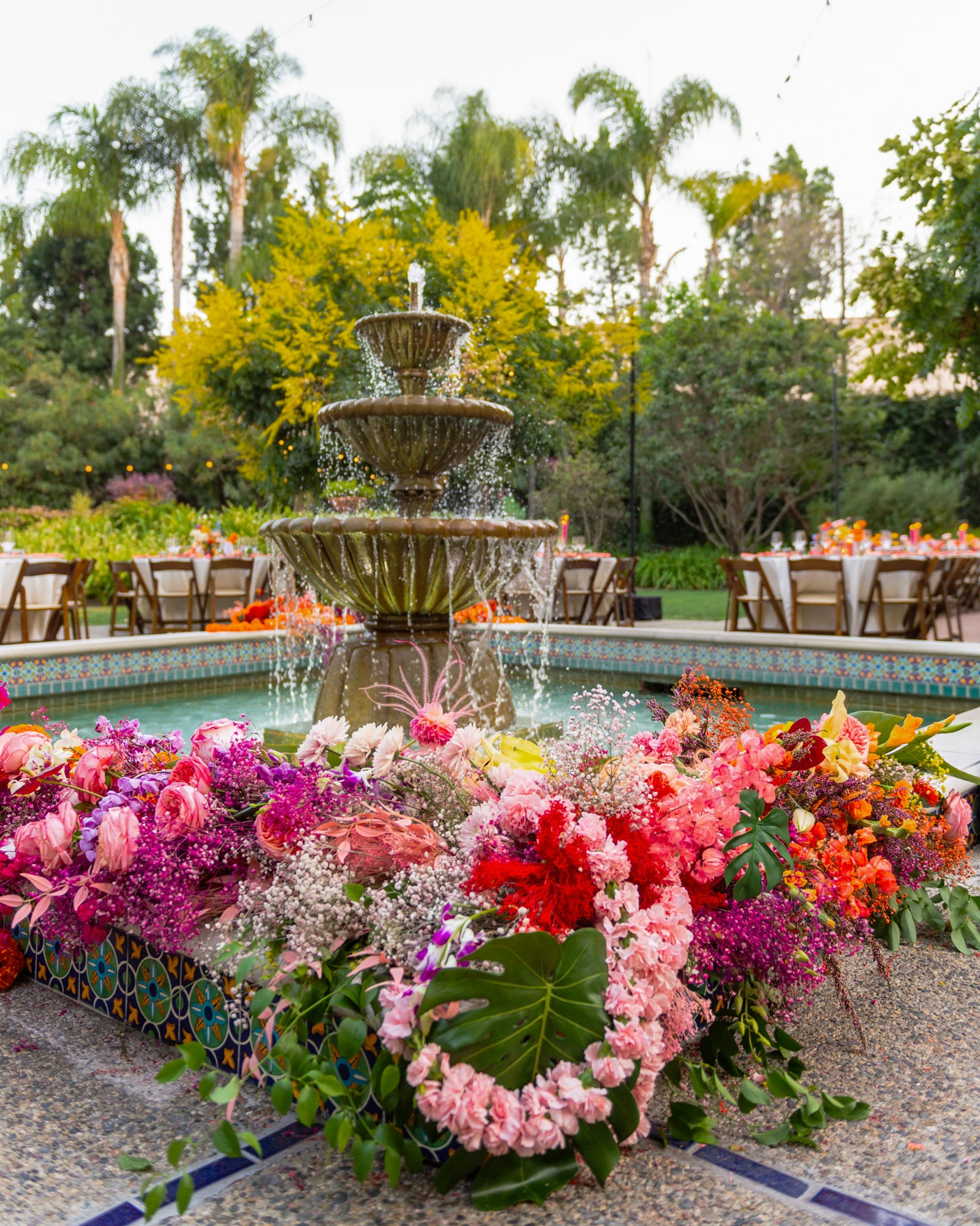 Wideshot of the multi-tiered water fountain, which is made of stone, with water gently cascading down from the top tier into the basin below. The fountain is surrounded by an abundance of vibrant, colorful flowers, including shades of pink, red, orange, purple, and white. Large tropical leaves, like monstera leaves, are interspersed among the flowers, adding texture and depth.