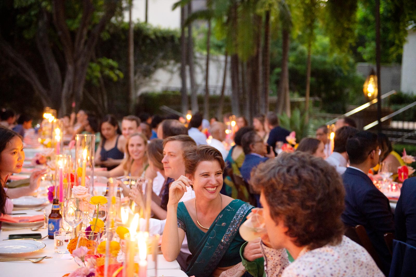Wide shot of guests sitting at a long, candlelit table outdoors in the evening. The atmosphere is warm and lively, with people chatting and smiling. The table is decorated with bright flowers and glowing candles.