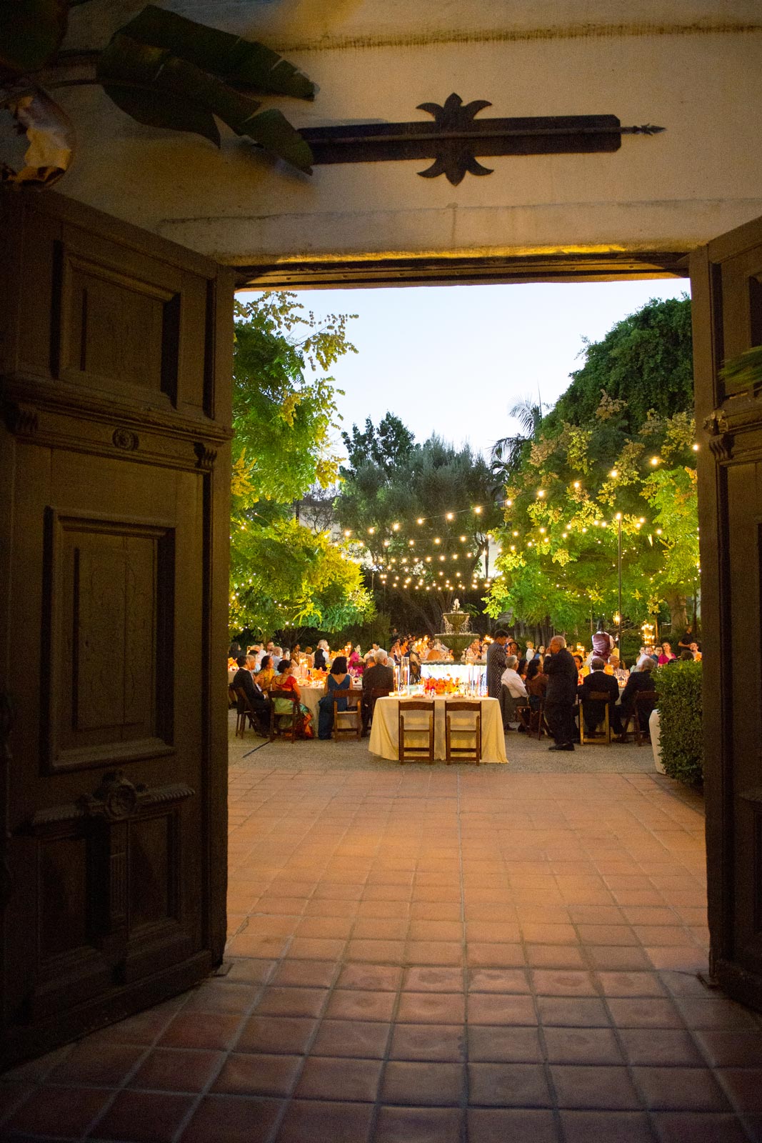 Wide shot in the evening during the reception viewed through an open doorway. String lights hang above the outdoor dining area, where guests are seated around tables. The fountain is visible in the center, and the setting is surrounded by greenery, creating a warm atmosphere.