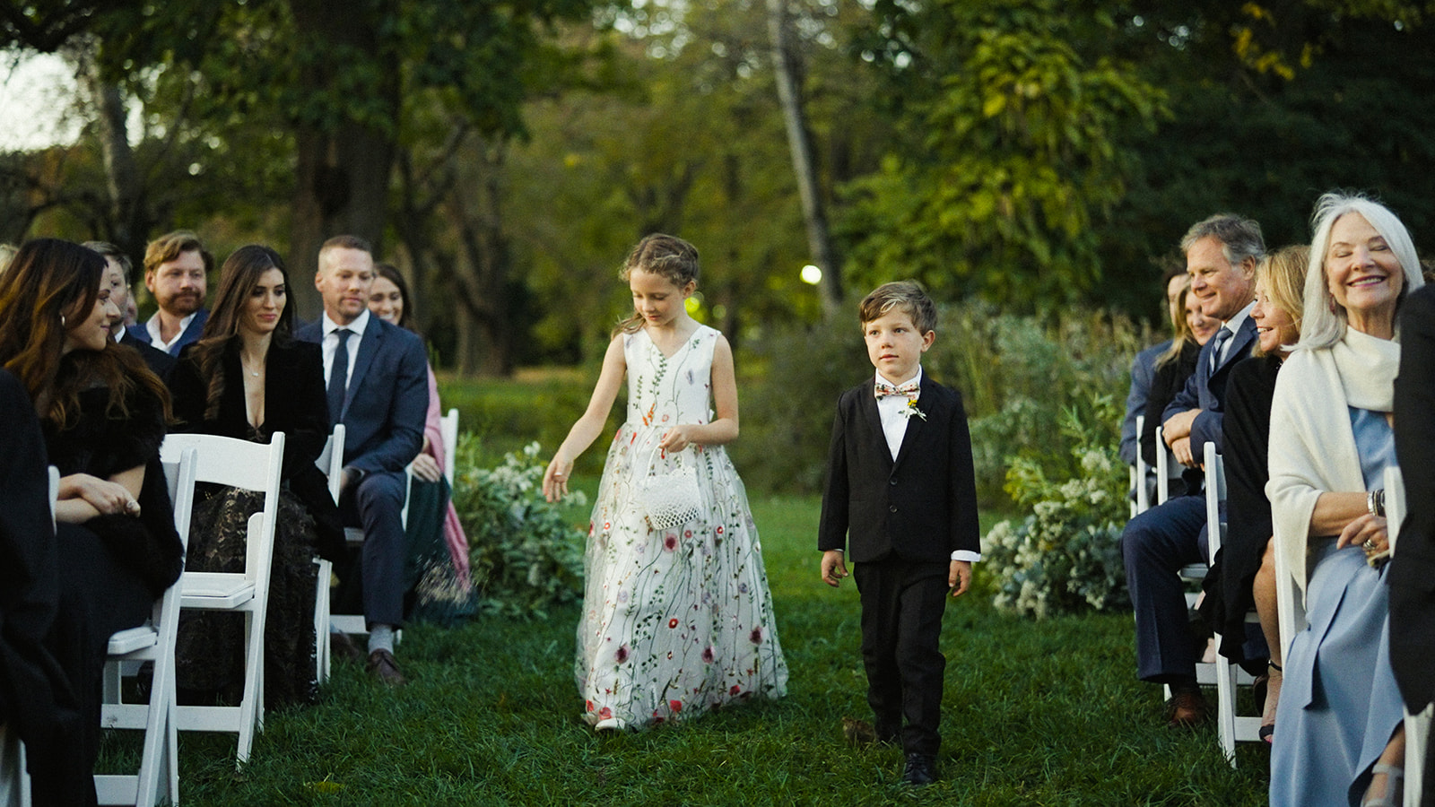 The flower girl and the ring bearer walk down a grassy aisle towards the camera. The girl on the left is wearing a long white dress decorated with embroidered flowers and carrying a small basket of flowers. The boy to her right is wearing a black suit and bow tie, looking serious as they walk. In the background, guests are seated on white chairs, smiling and watching them.