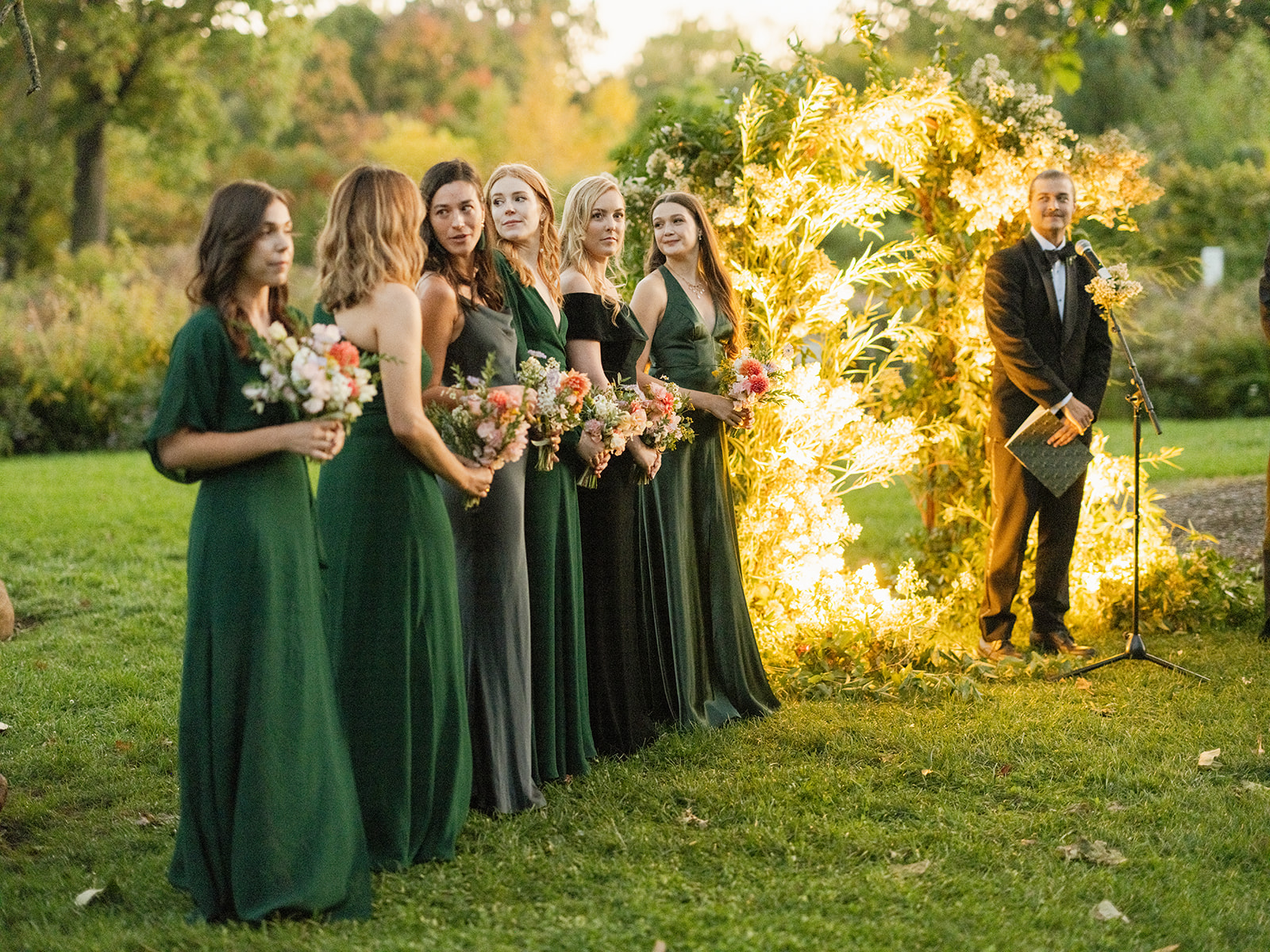 Wide shot of the bridesmaids standing in a line outdoors during the evening ceremony. Next to the arch, an officiant dressed in a tuxedo addresses the guests.