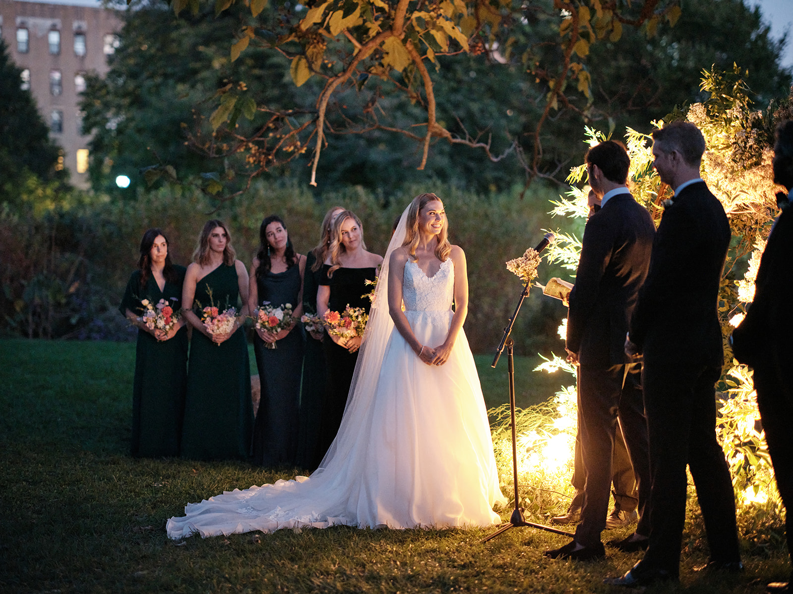 Wide shot of the ceremony with the focus on the bride gazing at the groom. The lighting from the arch adds a warm, magical glow to the scene, casting soft shadows. To the left, the bridesmaids stand in a row, holding bouquets and watching the ceremony.