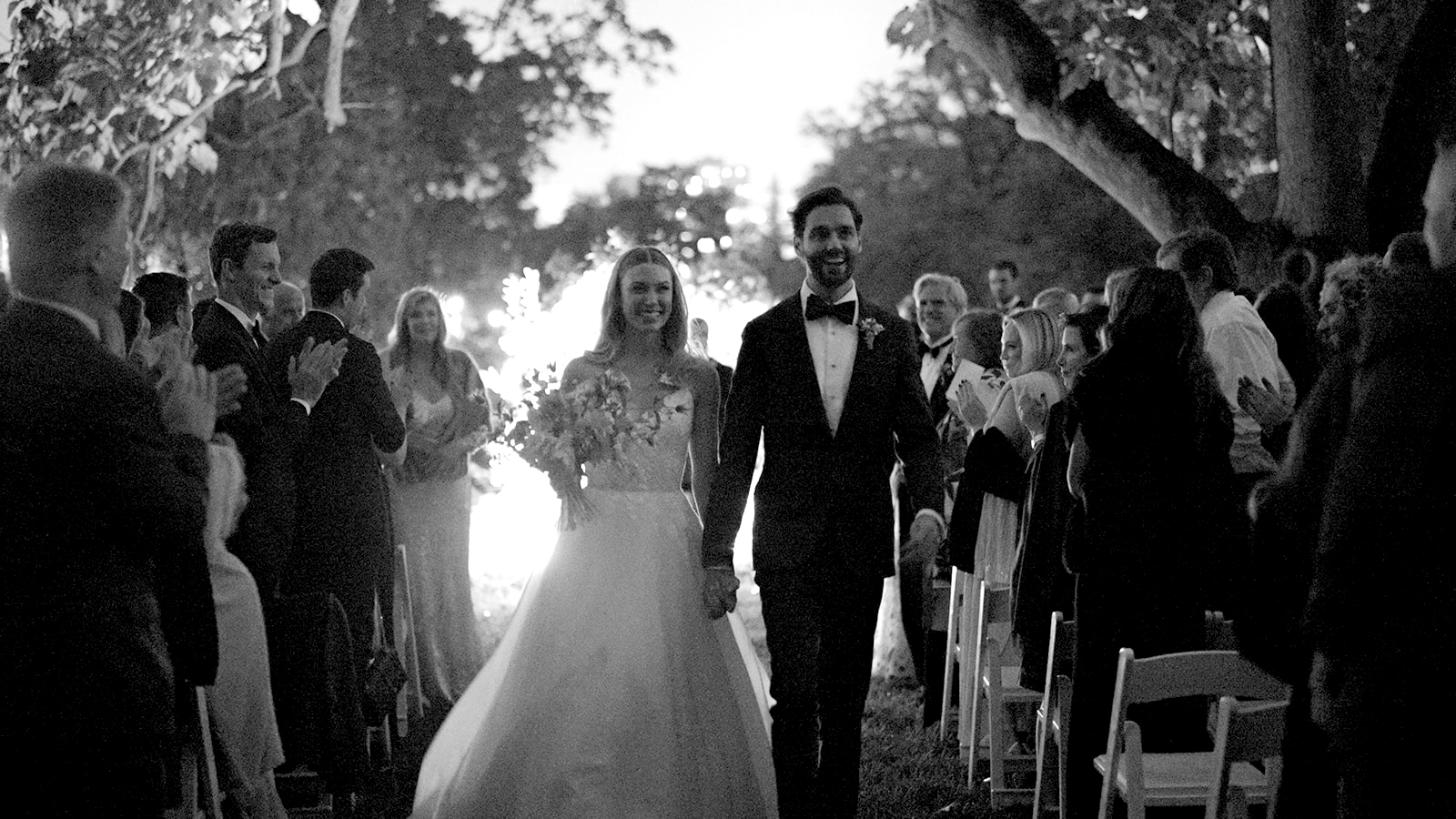 Black-and-white wide shot of the couple triumphantly walking towards the camera, now newlyweds, walking hand-in-hand down the aisle. The bride holds her bouquet with a serene smile while the groom beams joyfully. They are walking together as a married couple, framed by the soft light behind them.