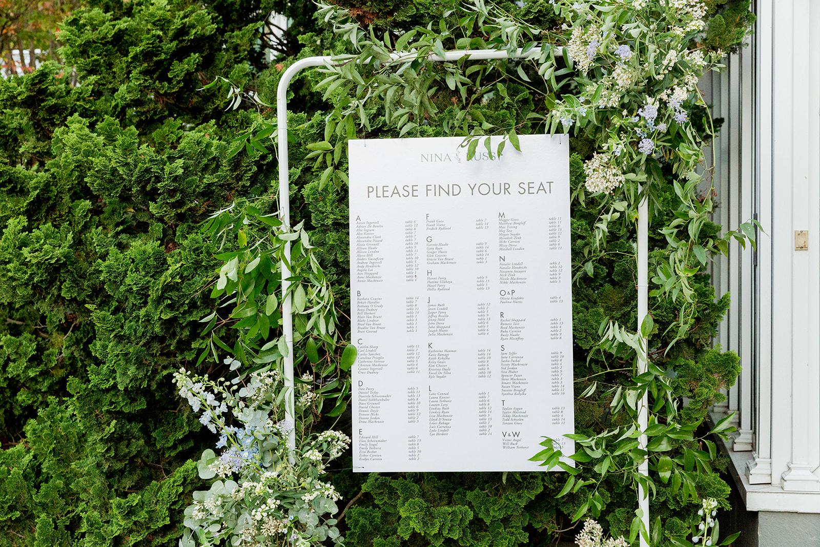 Medium shot of a large seating chart set outdoors against a lush green background of trees and foliage. The chart has a clean, minimalist design with the words "PLEASE FIND YOUR SEAT" in large letters at the top. Below this, the names of guests are listed in alphabetical order, along with their table assignments.