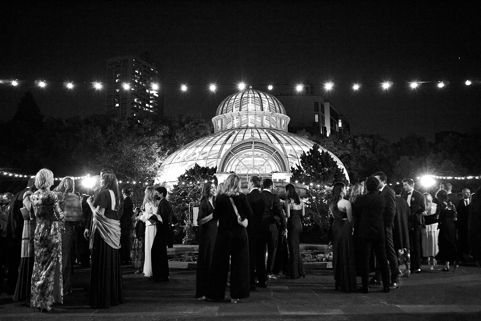 Black-and-white wide shot capturing the wedding reception outdoors at night, with a grand glass conservatory in the background, beautifully lit from within.