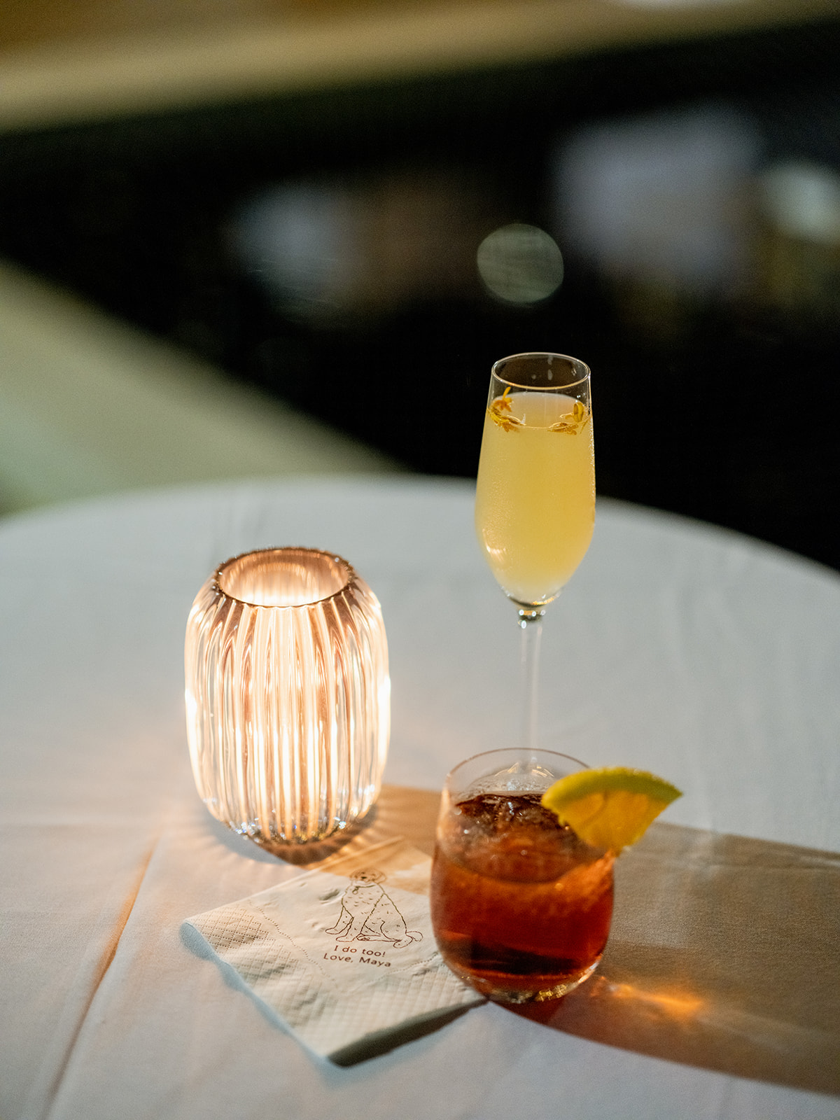 Close-up shot of a small round table with two signature cocktails created just for the wedding. On the left, a warm glowing candle holder softly illuminates the scene. The drink on the left is in a tall flute (the French 85) filled with a light Gin and St. Germaine-focused golden-yellow drink, topped off with Prosecco and garnished with small delicate flowers. The drink on the right is in a short, round glass filled with Gin and Campari garnished with an orange slice entitled the “Ninagroni.” Below the glasses, a napkin features a printed illustration of a dog with the words "I do too! Love, Maya."