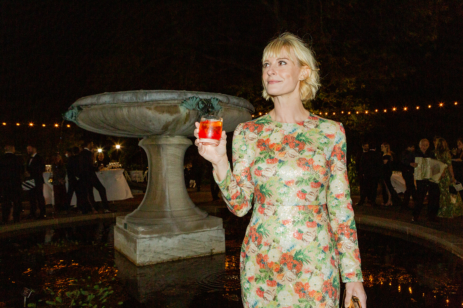 A party guest with a patterned, sequined gown standing by a large, elegant stone fountain at night. She holds a bright red cocktail in her hand and looks off to the side calmly and composedly. The background is dimly lit, with string lights creating a warm glow above, and guests can be seen mingling in the distance.