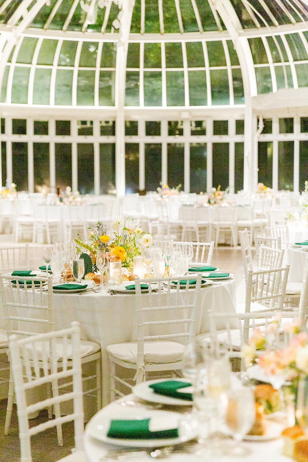 Wide shot of the wedding reception inside a grand, glass-domed venue, like a conservatory. The room is filled with round tables covered in white linens, each adorned with elegant centerpieces of flowers in soft colors like yellow, white, and orange. The floral arrangements are complemented by glassware, plates, and candles, creating a warm, inviting atmosphere. White chairs surround the tables, and neatly folded green napkins are placed on each plate. In the background, large glass windows reach up to the ceiling, giving the space an open and airy feel.