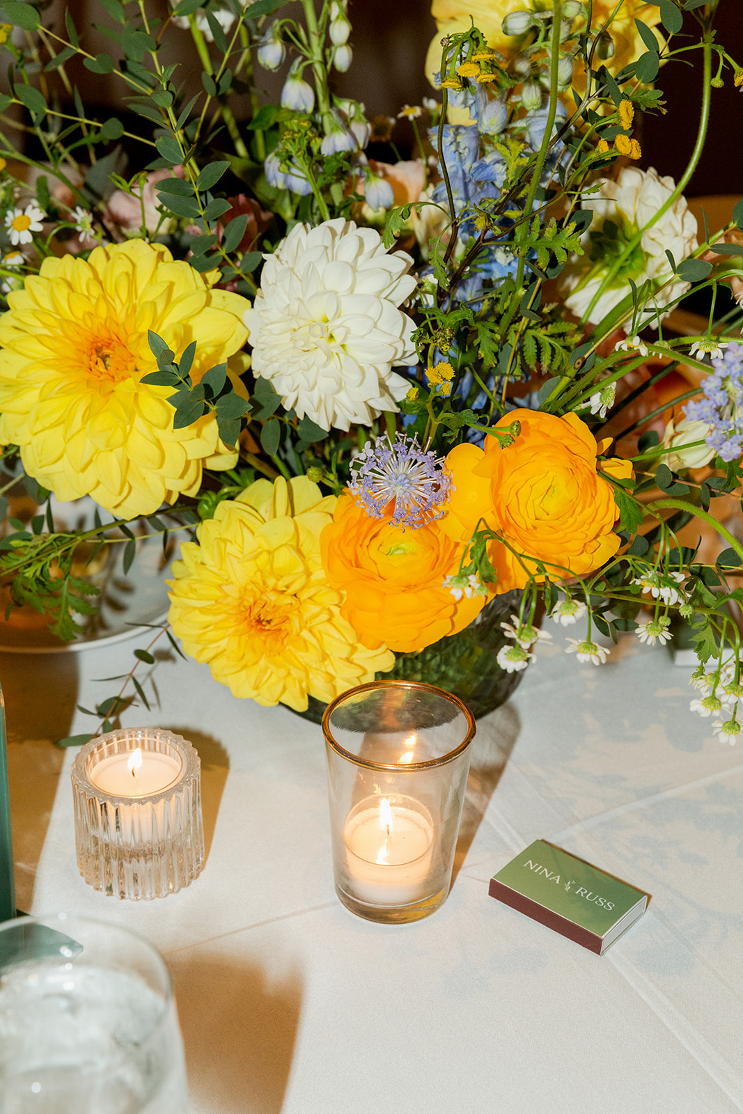 Close-up of a tablescape with an on-camera flash of a beautiful floral arrangement on a table featuring vibrant yellow and white flowers with some small purple and blue accents. Surrounding the flowers are small Diptyque votive candles, casting a warm glow across the table. A matchbox named "Nina & Russ" is also on the table.