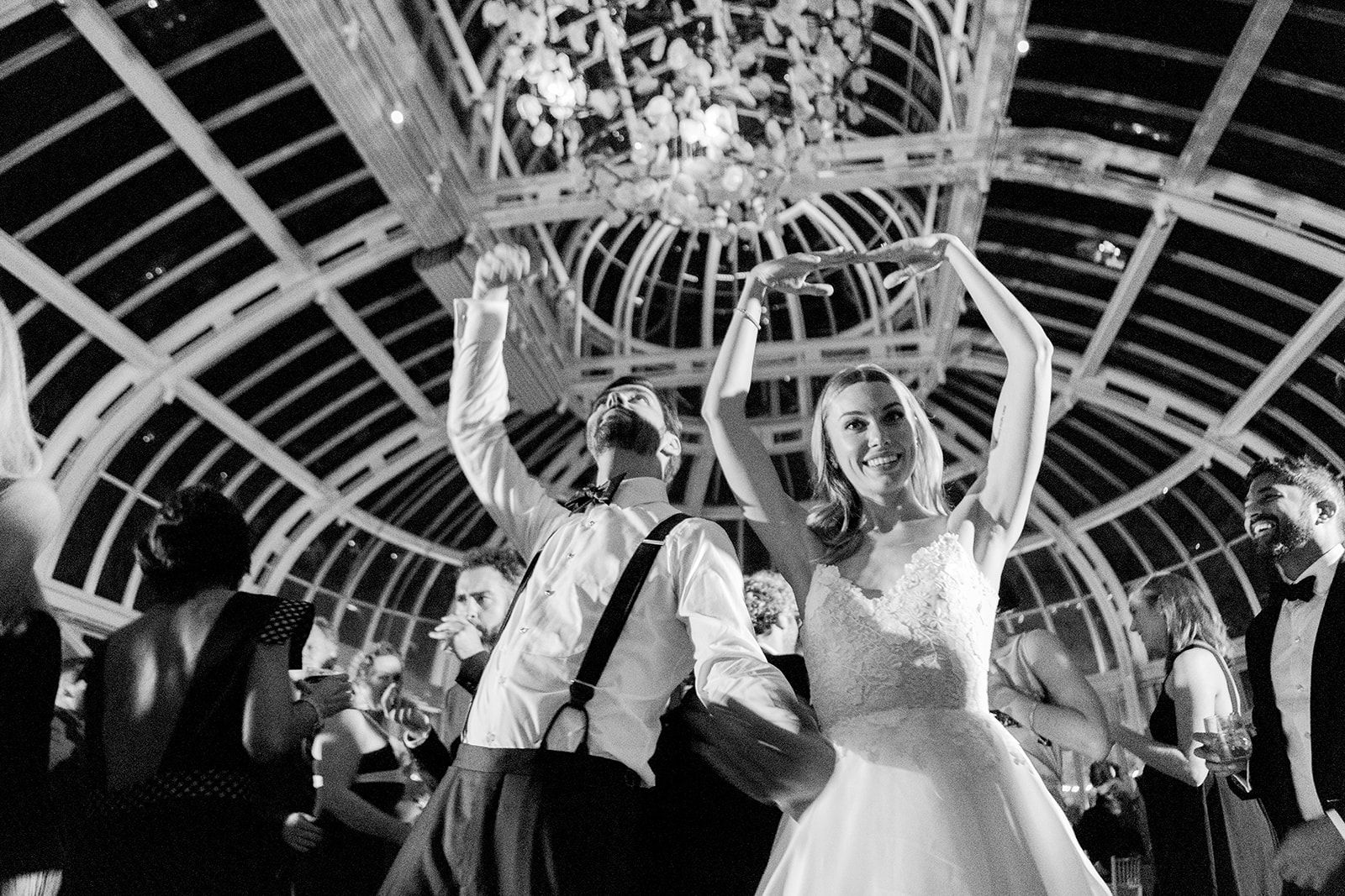 Candid black and white photo of a joyful and lighthearted bride and groom dancing and celebrating under the grand glass ceiling. Still full of energy, the bride and groom lead the dance floor in a fun, spirited movement, surrounded by their guests.