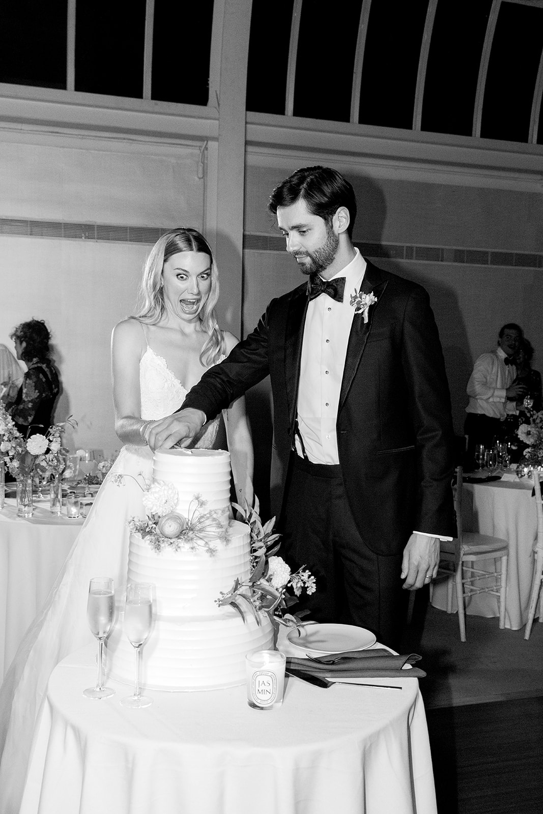 Black-and-white capture of the bride and groom cutting their wedding cake. The bride displays a playful, exaggerated expression of excitement or surprise, adding a lighthearted tone to the moment, while the groom remains focused on the task of cutting the cake. The multi-tiered cake is elegantly decorated with flowers, and two champagne flutes are set on the table before them.